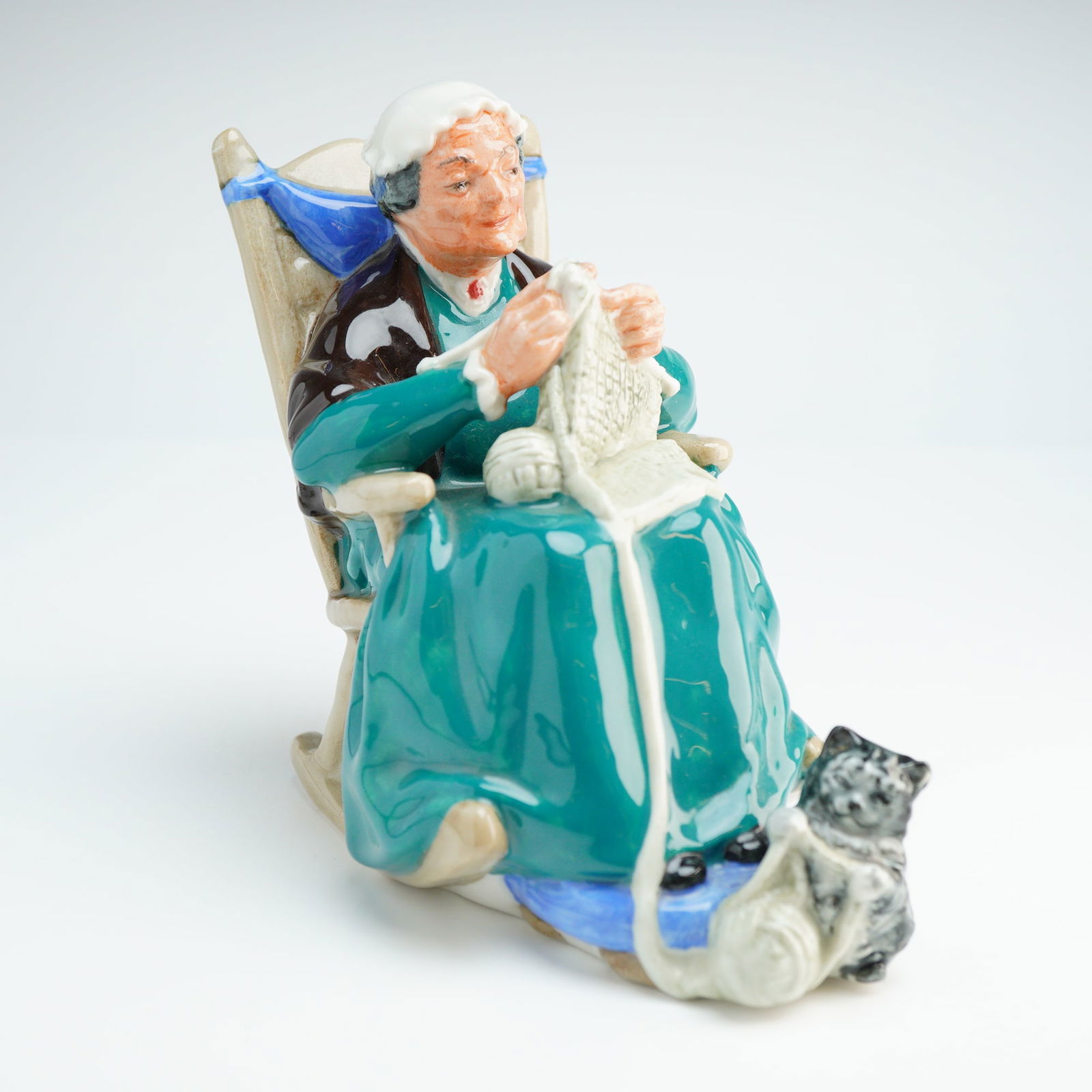 Royal Doulton Figurine Twilight HN2256: The piece depicts an elderly woman seated in a tan rocking chair, wearing a teal dress, a brown shawl, and a white cap. She is shown knitting with white yarn, while a small cat sits at her feet. The r