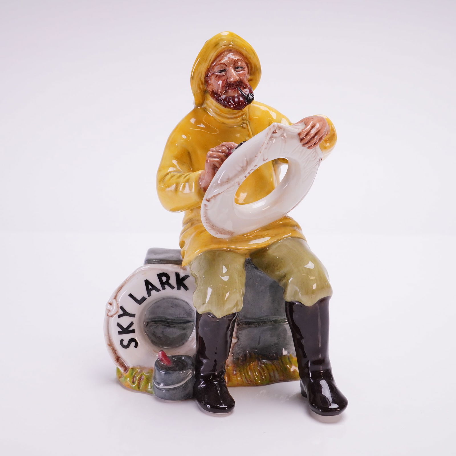 Royal Doulton Figurine, Boatman Skylark HN2417: The piece depicts a seated male figure in traditional maritime attire, including a yellow sou'wester, yellow raincoat, and black boots. The figure is portrayed smoking a pipe and holding a white lifeb