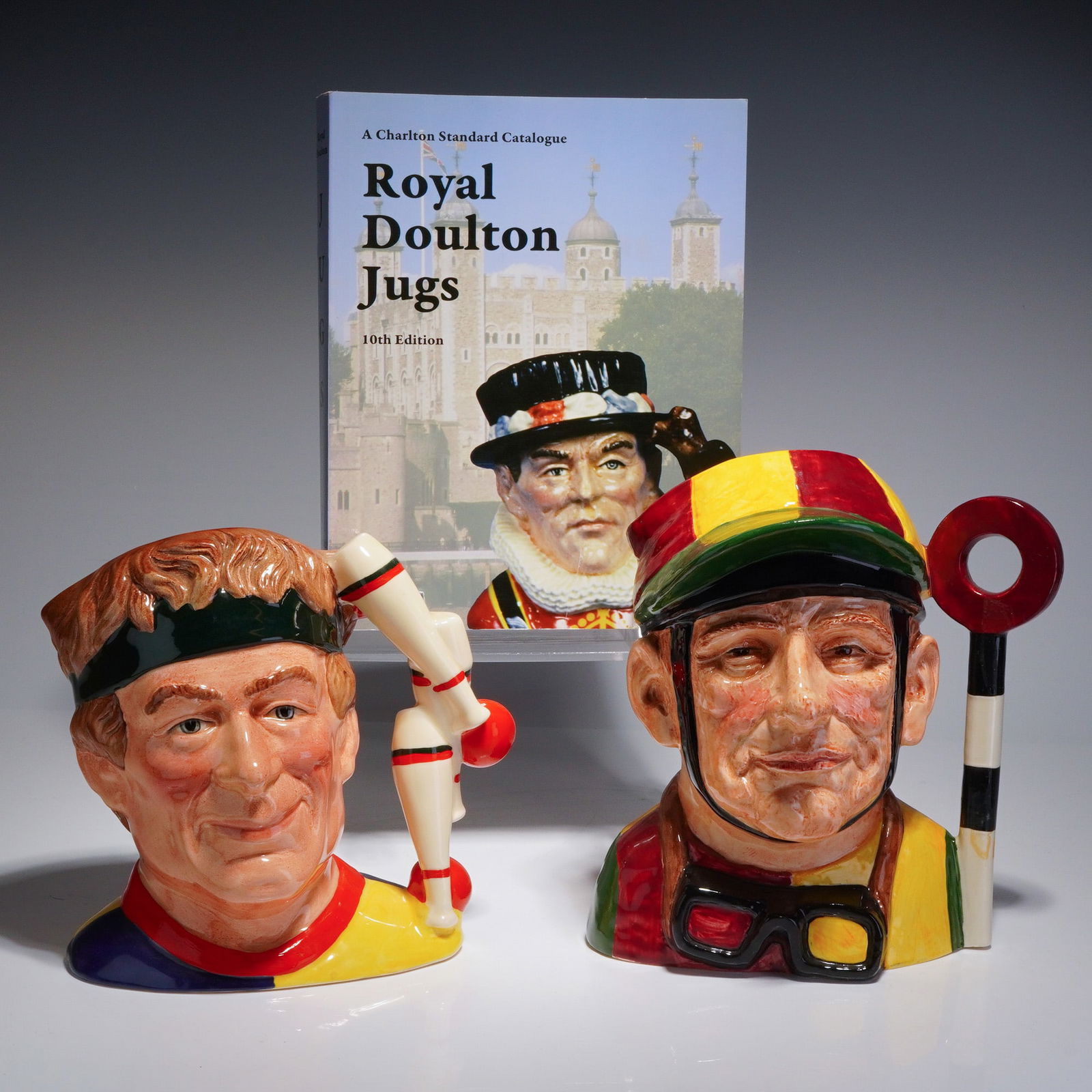 3pc Royal Doulton Large Character Jugs and Jugs Reference Book: A collection of two Royal Doulton ceramic character jugs and a reference book. The group includes the large-sized character jug The Juggler, model D6835, designed by Stanley James Taylor and dated 198