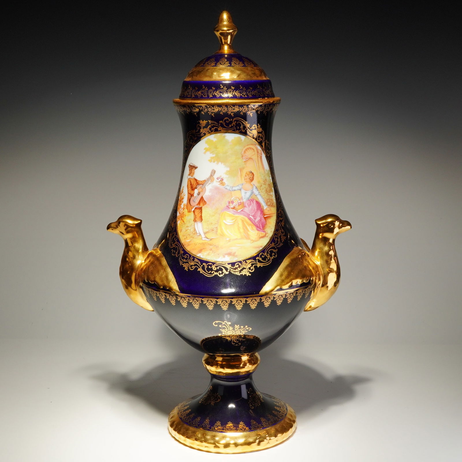 Limoges Rehausse Main Lidded Vase, Napoleon: A porcelain lidded pedestal vase featuring a cobalt blue ground with extensive raised gilt decoration, the body centered with an oval hand painted courting scene after Fragonard depicting a seated lad