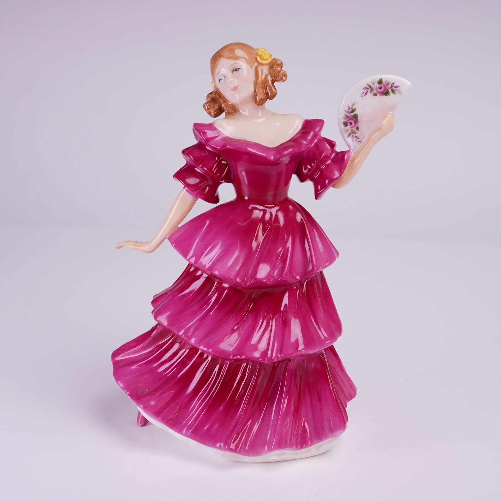 Royal Doulton Figurine, Jennifer HN3447: The piece depicts a woman modeled in a romantic, traditional style, wearing a tiered dress rendered in a vibrant magenta or fuchsia color. The figure holds a floral-decorated fan in her right hand. Th