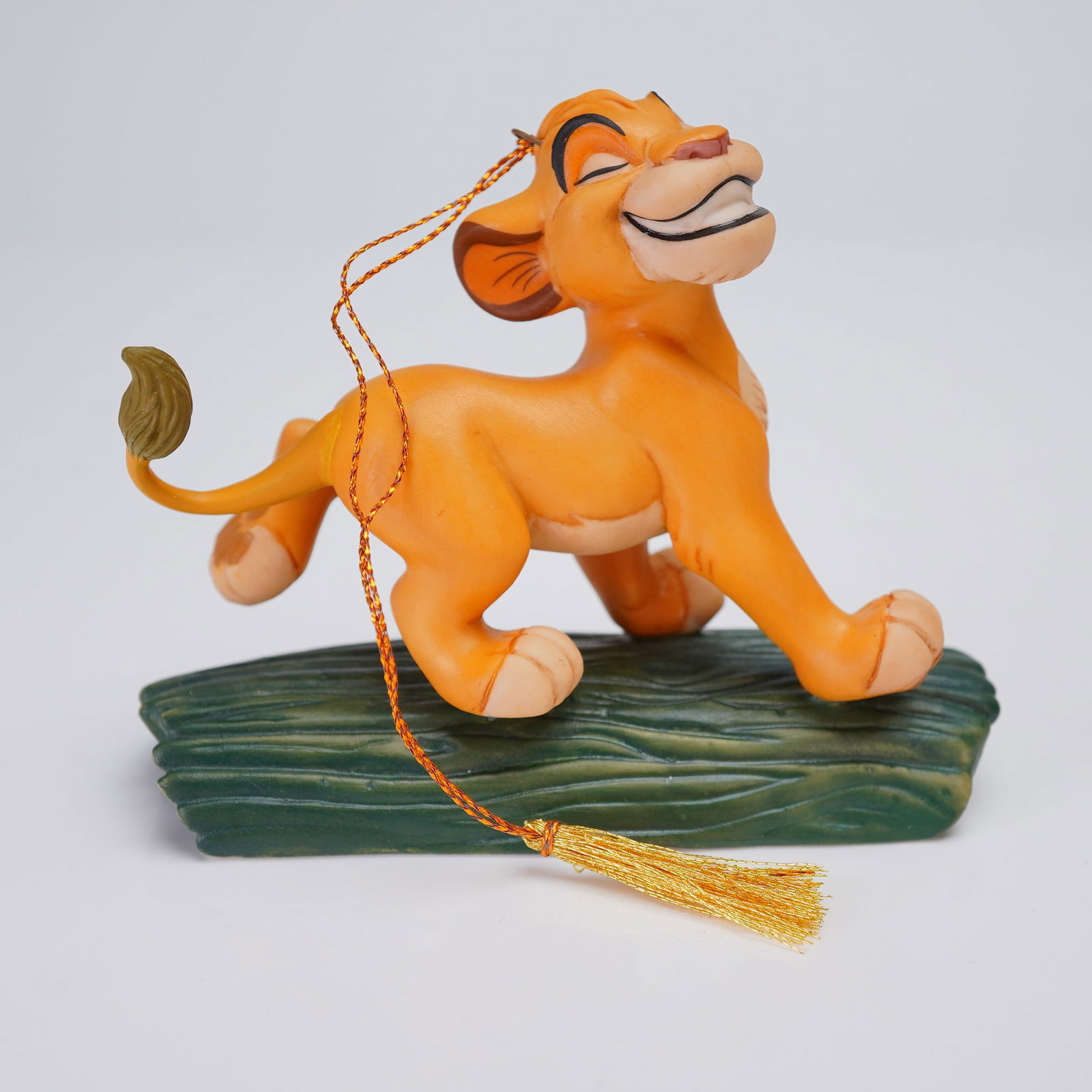 Walt Disney Ornament, The Lion King Hakuna Matata Simba: The figurine is executed in painted ceramic or resin, showing Simba standing in a playful pose atop a textured dark green log base. The piece is designed as a hanging ornament, featuring an attached o