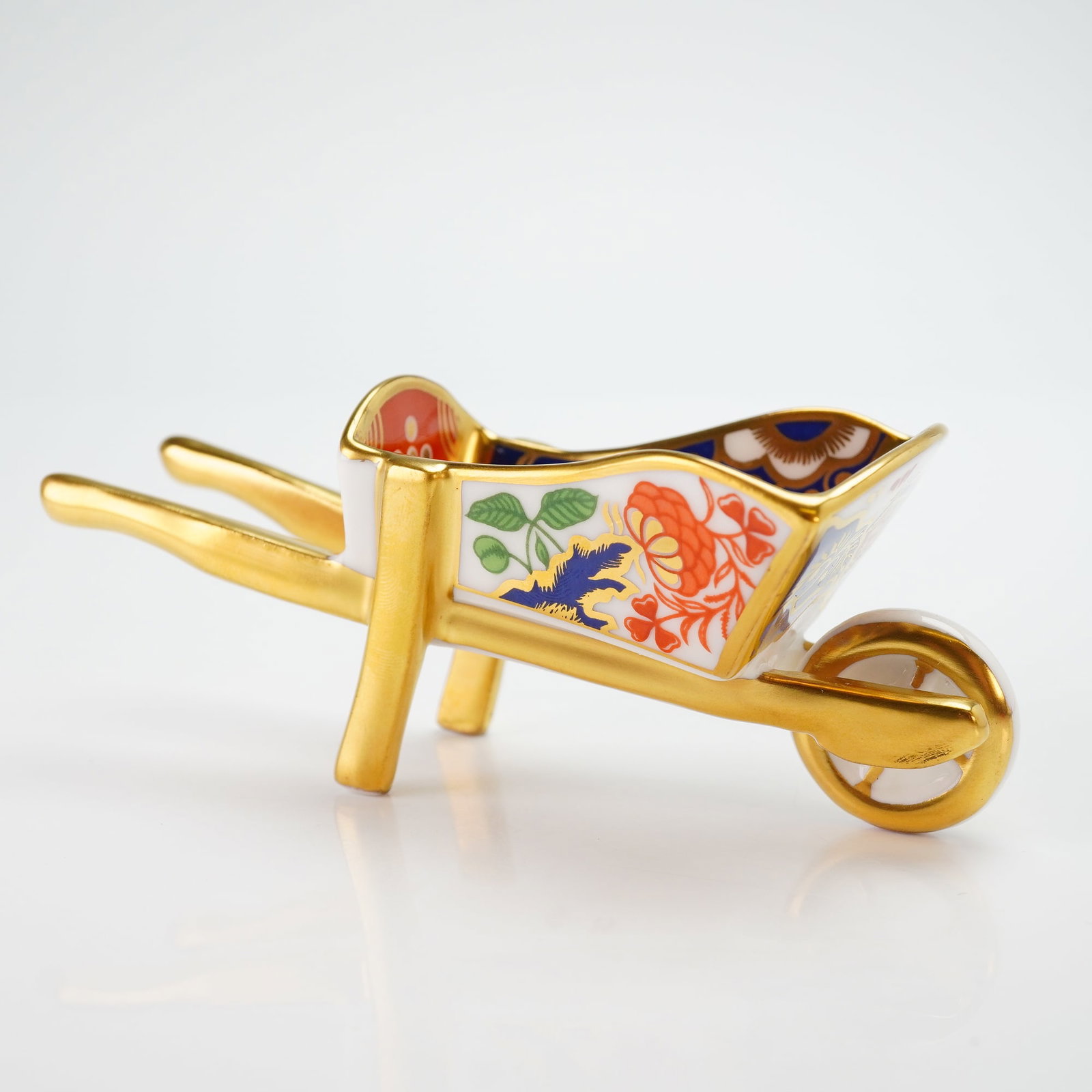 Royal Crown Derby Miniature Wheelbarrow: This piece is constructed of fine bone china and decorated in a traditional Imari style featuring cobalt blue, iron red, and gold gilt accents with floral and scroll motifs. The handles, support legs,