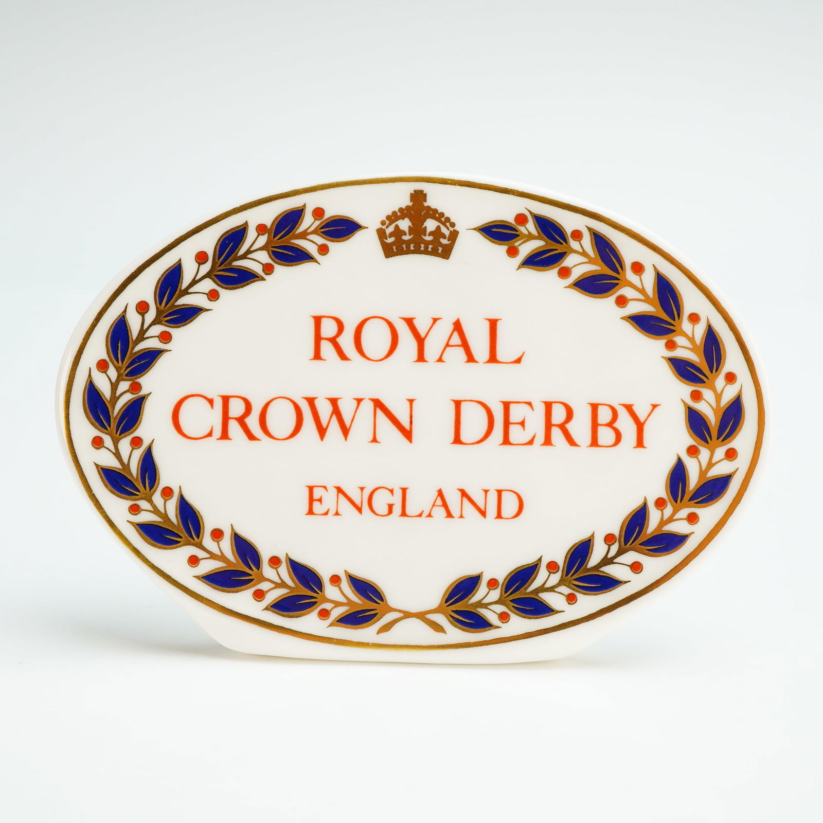 Royal Crown Derby Dealer Display Name Plaque: A Royal Crown Derby English fine bone china dealer display name plaque of oval form with an integrated flat base. The front is decorated with the brand name ROYAL CROWN DERBY ENGLAND in orange script,