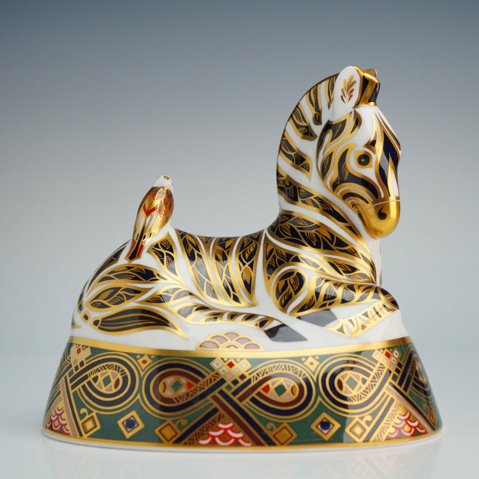 Royal Crown Derby Bone China Zebra Paperweight: A Royal Crown Derby porcelain paperweight figurine depicting a recumbent zebra with a bird perched on its flank. The piece is constructed of English bone china and decorated in a stylized Imari-inspir