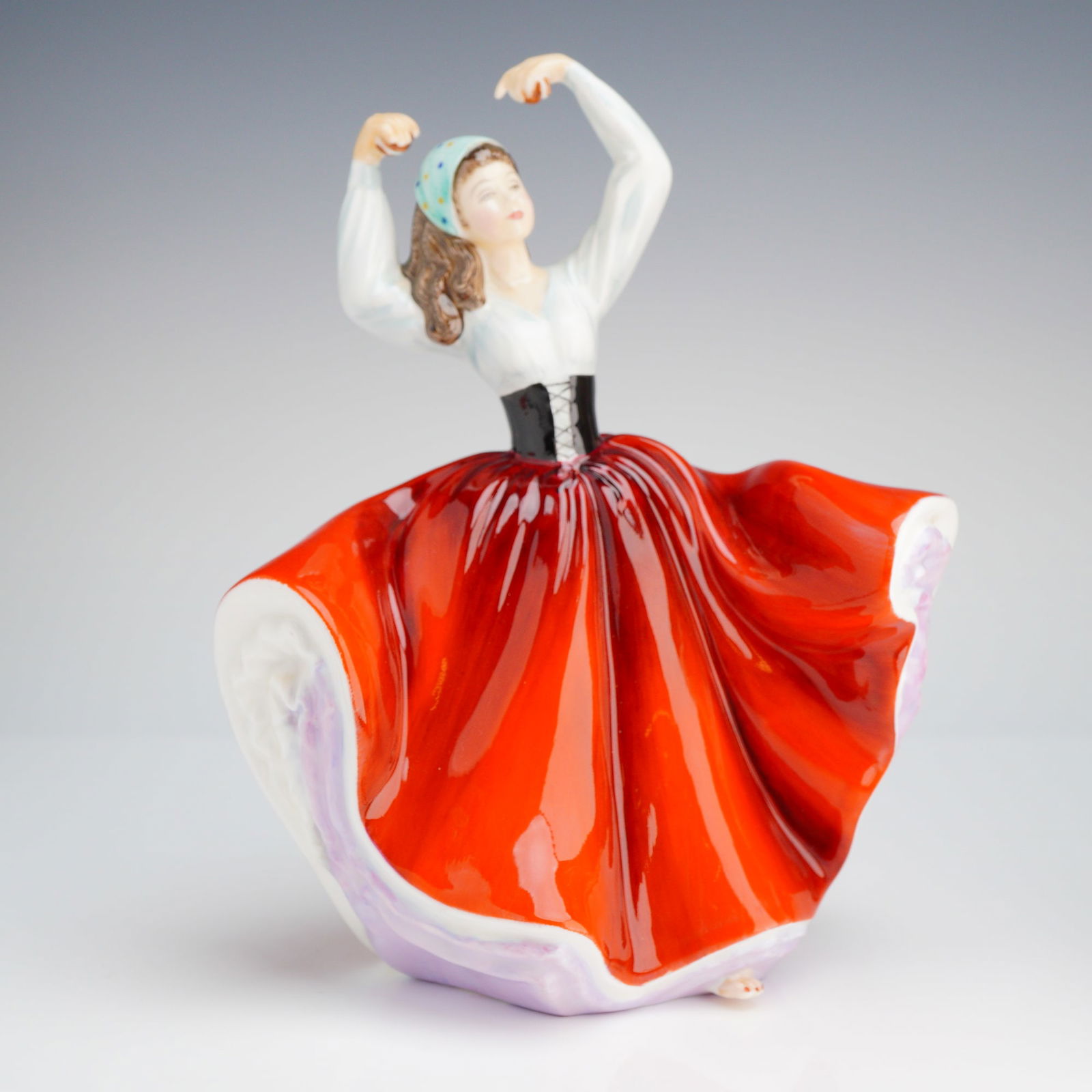 Royal Doulton Figurine, Karen HN2388: The figure is modeled as a female dancer in a dynamic, swirling pose with both arms raised above her head. She is dressed in a white peasant blouse, a black lace-up bodice, and a vibrant red skirt tha