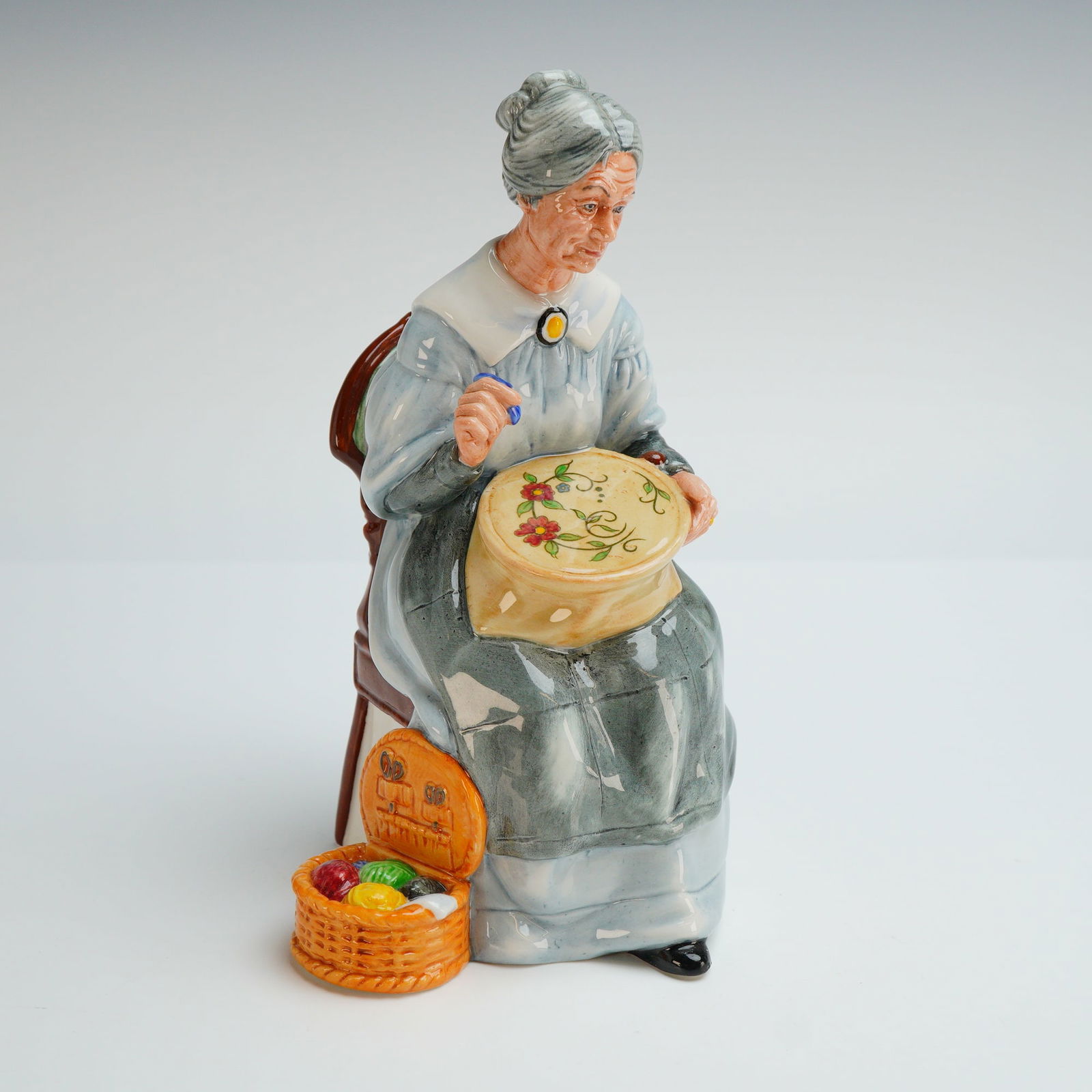 Royal Doulton Figurine, Embroidering HN2855: The figure depicts an elderly woman with grey hair styled in a bun, seated on a brown high-backed chair featuring green upholstery. She is dressed in a pale blue gown with a white collar and a dark gr