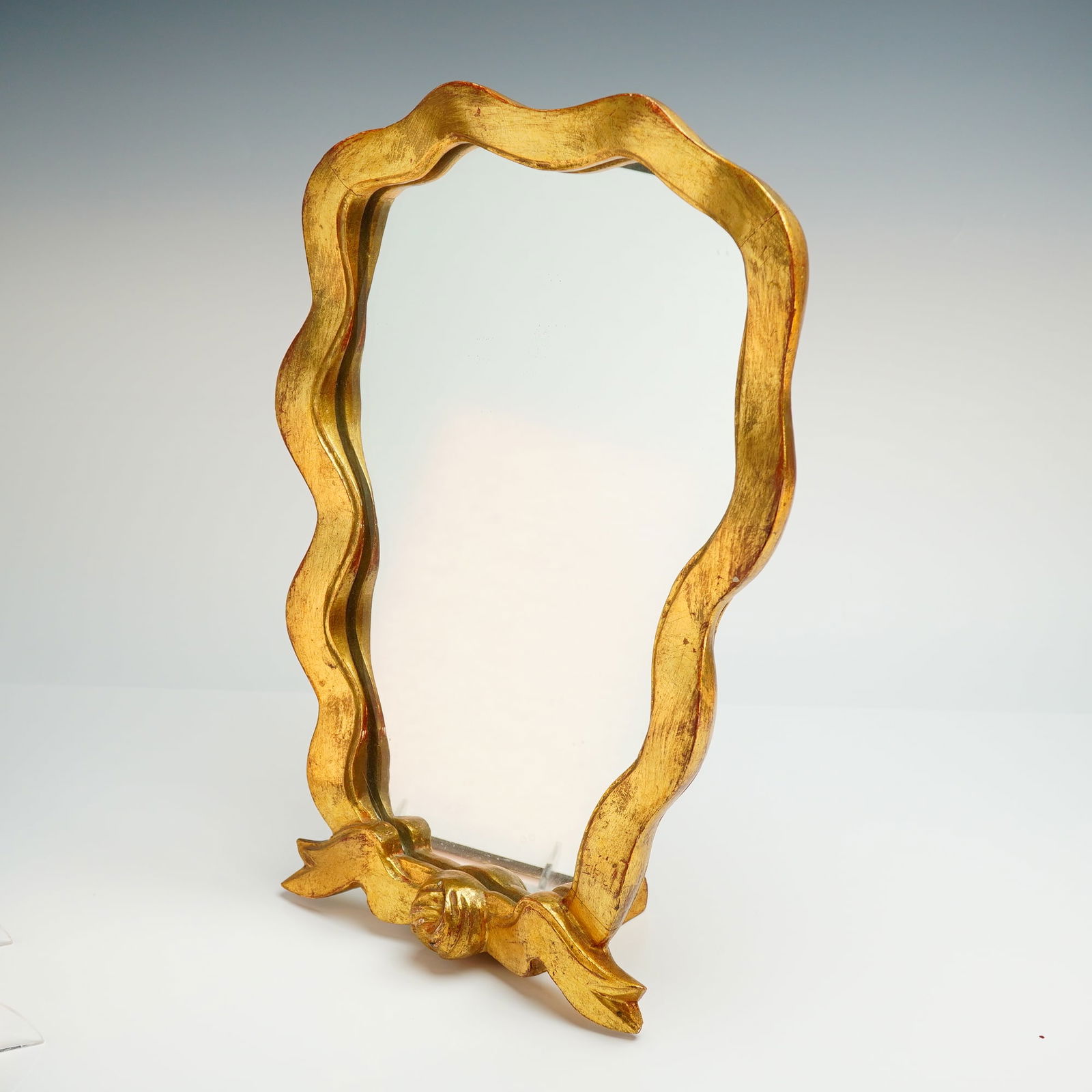 Italian Hollywood Regency Gilt Wood Table Mirror: This mid-20th century Italian Hollywood Regenct gilt wood table mirror features a wavy, organic-shaped frame finished in gold leaf over a red bole undercoat. The decorative base is characterized by a