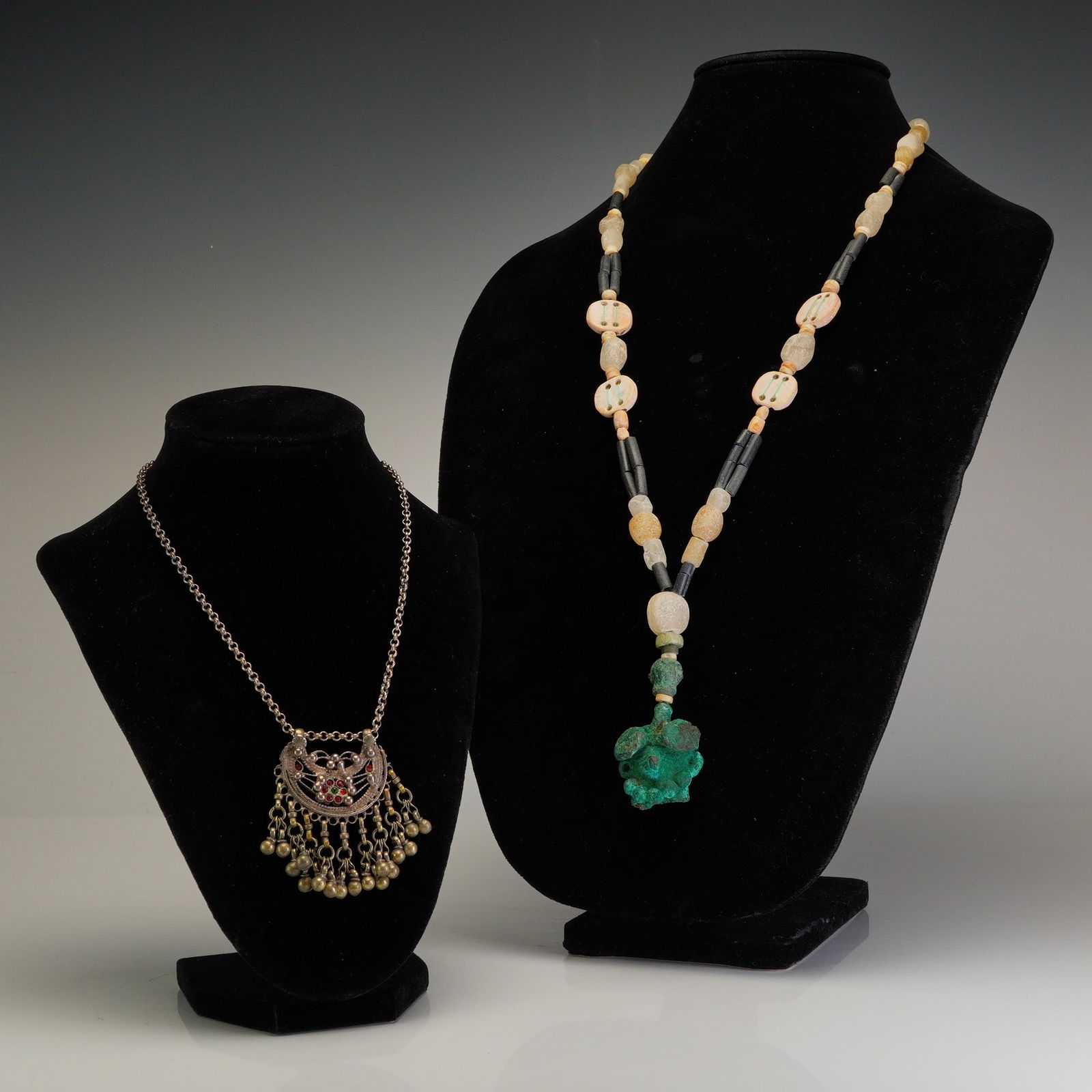 2pc Pendant Necklaces with Mixed Beads and Silver-Tone Metal: This lot features two ethnographic-style necklaces. The first necklace has a silver-tone metal crescent-shaped pendant with intricate filigree ornamentation, set with small red and green cabochons, an