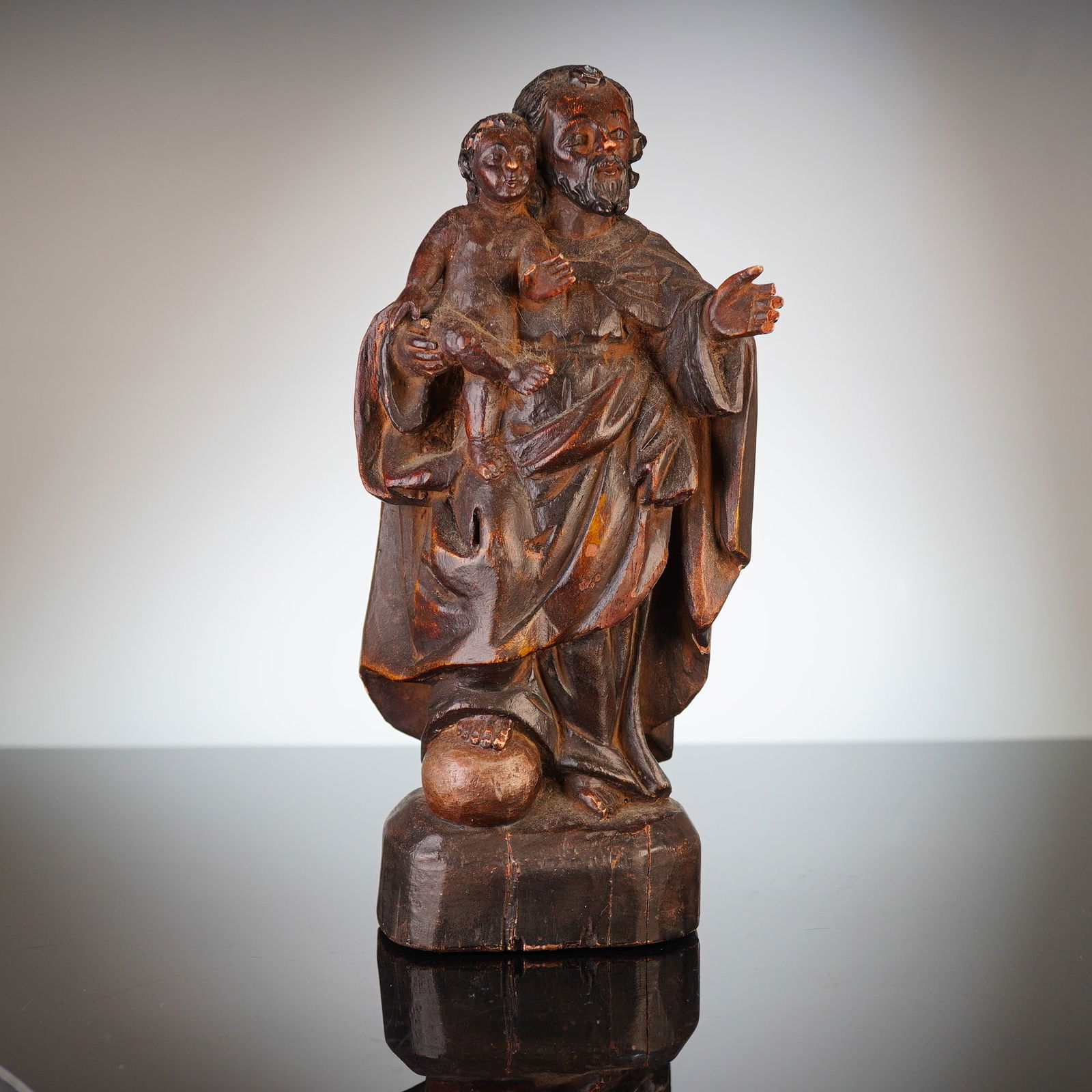 Carved Wooden Figurine of Saint Joseph Holding the Baby Jesus: A carved wooden figurine of Saint Joseph holding the baby Jesus, rendered in a colonial or folk art style. The sculpture is crafted from a dark-toned wood featuring a dark brown patina with visible