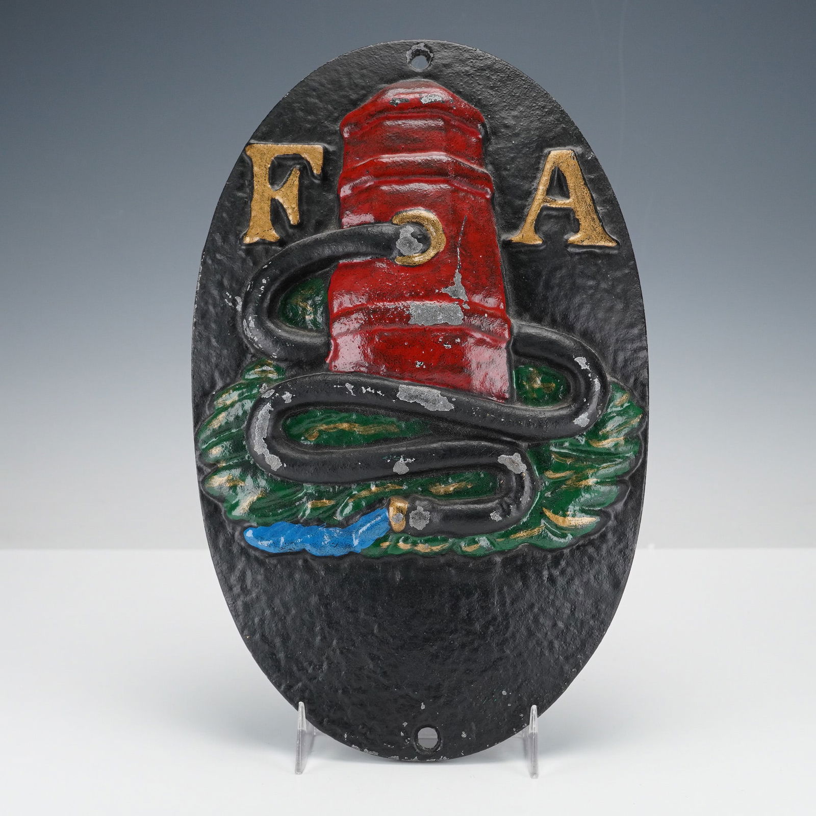 Fire Association of Philadelphia Cast Iron Fire Mark Plaque: This oval cast iron plaque shows a raised red hydrant wrapped with a black hose above green grass with a stream of blue water shooting out, with the gold colored letters F and A at the top. It has a