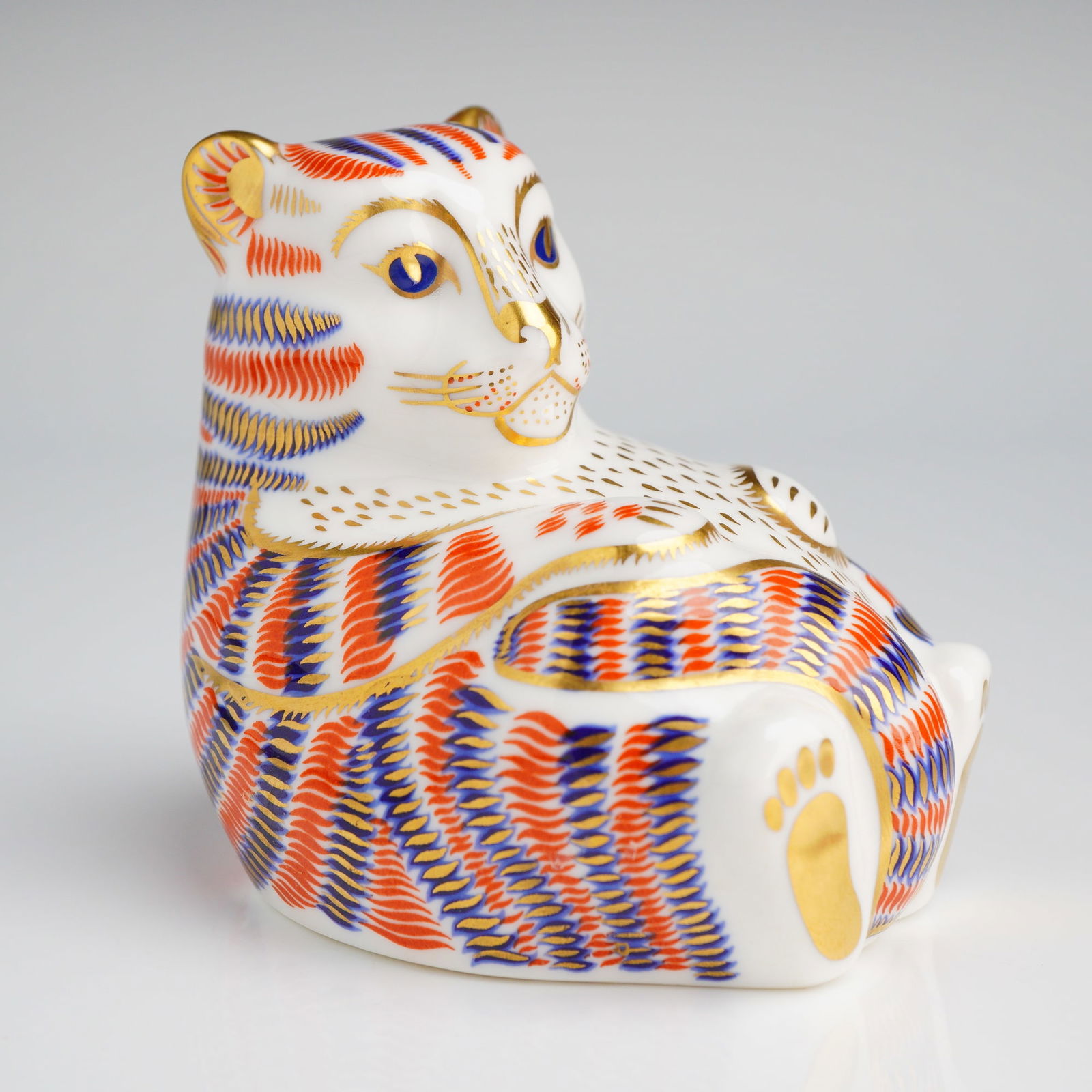 Royal Crown Derby Tiger Cub Paperweight: A Royal Crown Derby English bone china paperweight depicting a seated tiger cub. The figurine is decorated in an Imari-style palette featuring iron-red, cobalt blue, and gold gilding over a white glaz