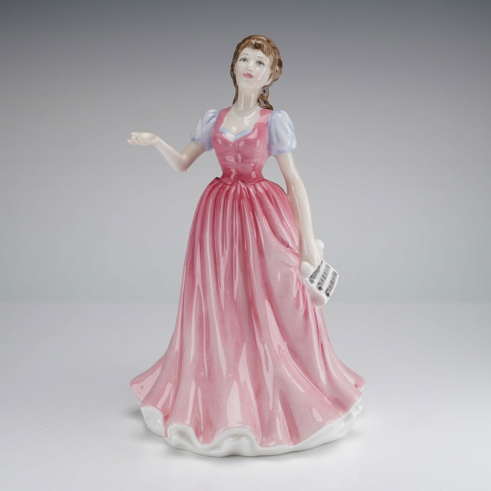 Royal Doulton Figurine Sweet Music HN 4302: The figurine is styled as a young woman in a long, flowing, glossy pink dress detailed with light blue puff sleeves. The figure is posed with her right hand extended slightly and holds a rolled sheet