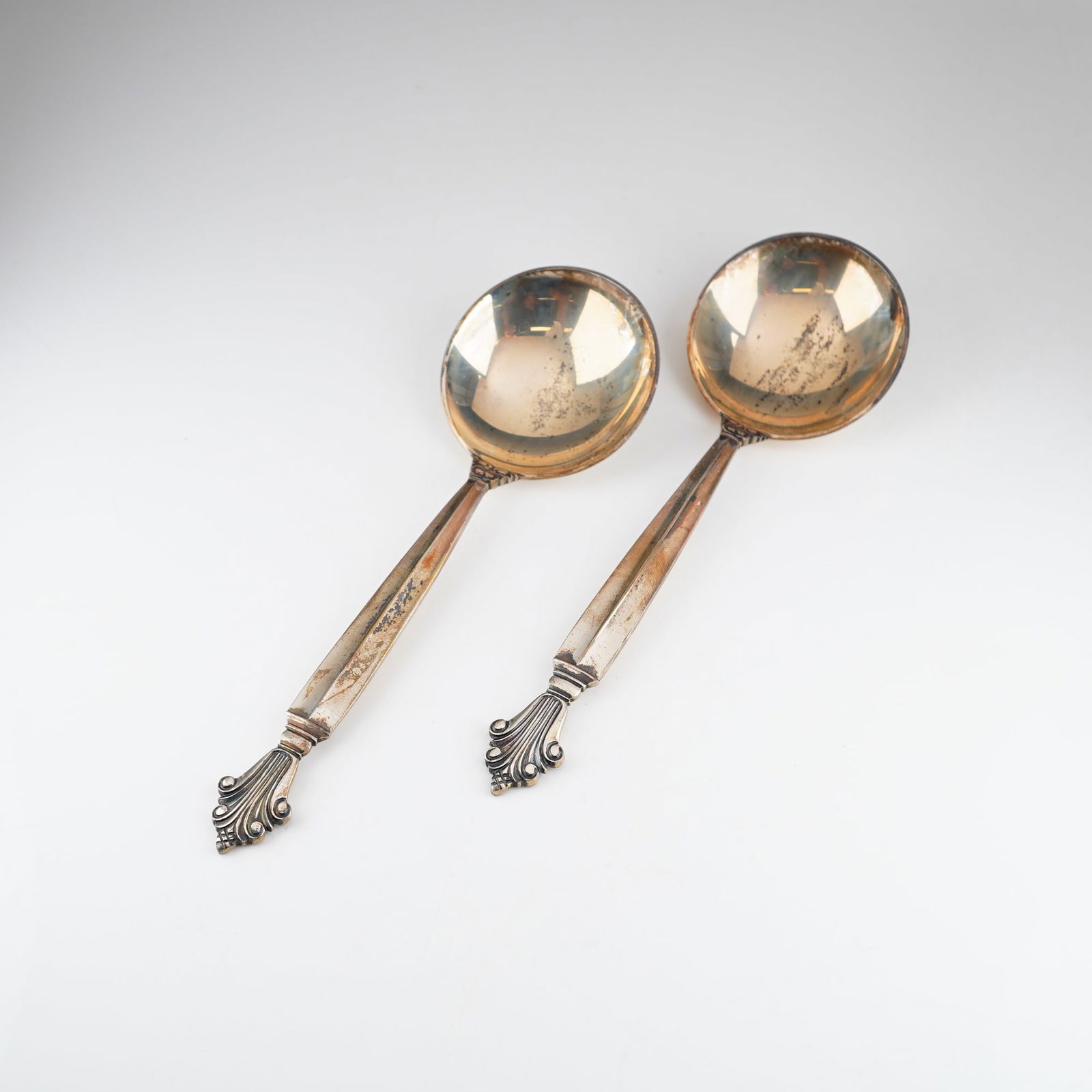 2pc Georg Jensen Sterling Silver Round Bouillon Soup Spoons, Acanthus: A pair of Danish sterling silver serving spoons in the Acanthus pattern. Each spoon features a large circular bowl and a tapered, faceted handle. The handles terminate in a decorative, stylized