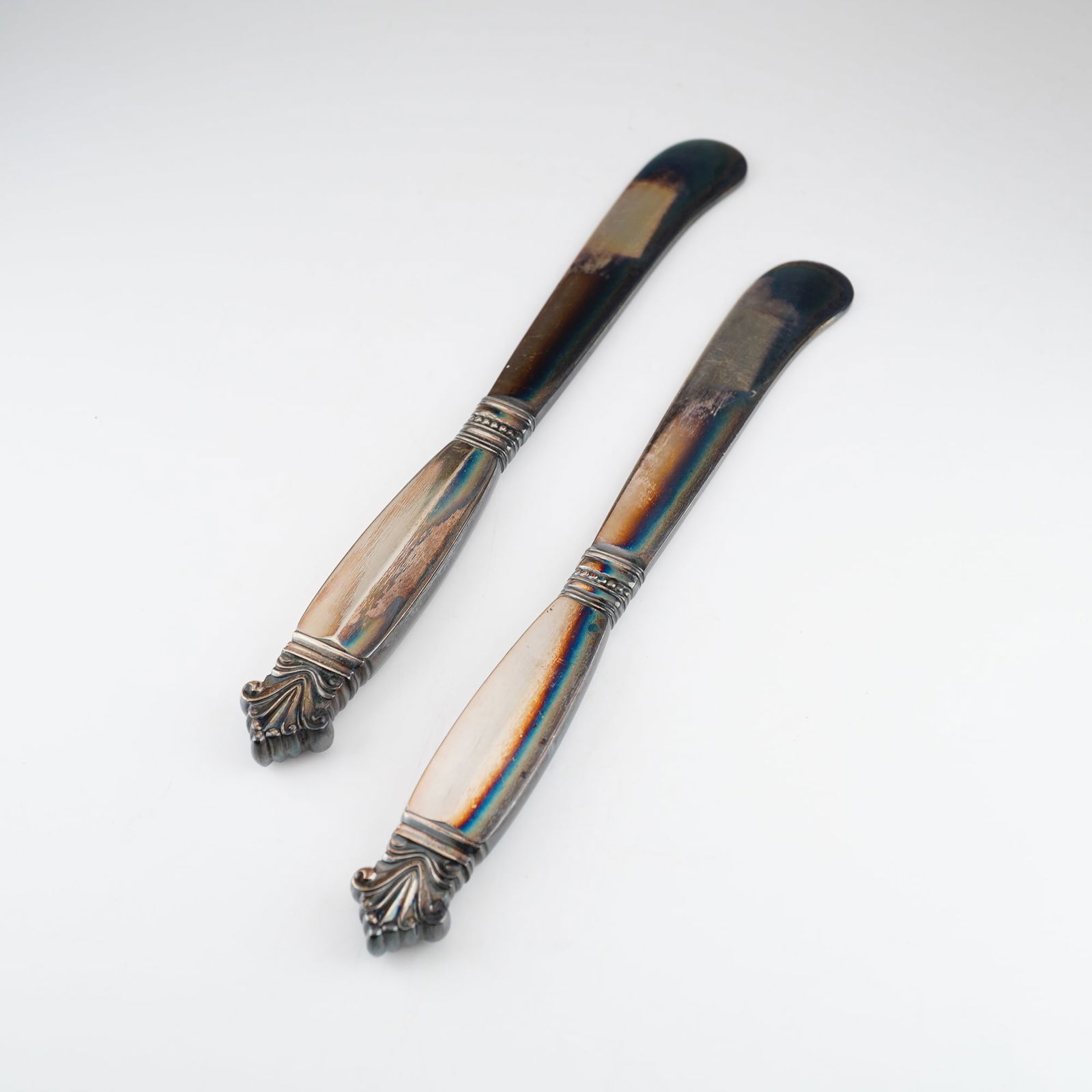 2pc Georg Jensen Sterling Silver Butter Spreaders, Acanthus: Each piece features a handle with a stylized acorn finial and decorative beaded bands, tapering to a broad, curved spreading blade. The items are marked with the Georg Jensen dotted oval hallmark and