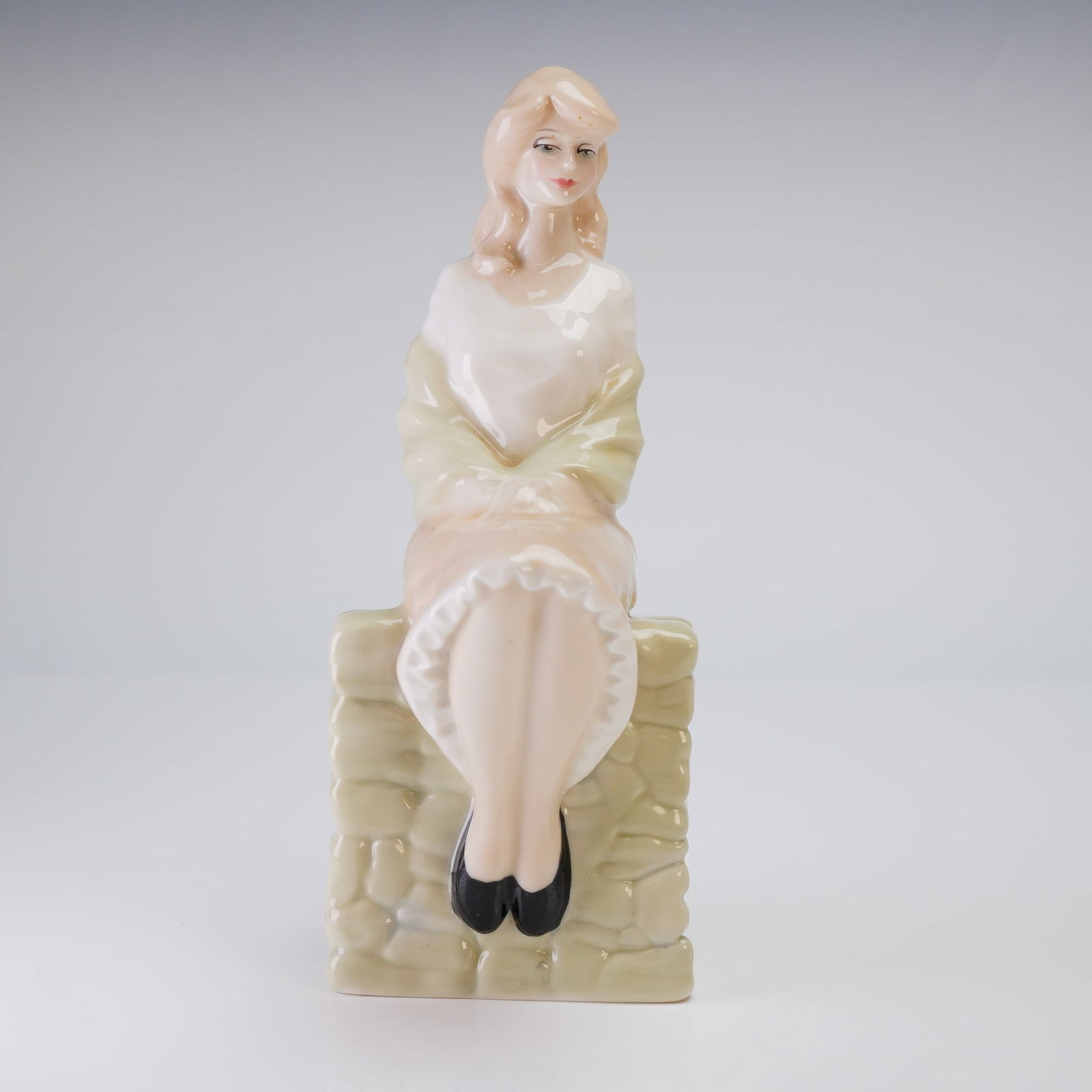 Royal Doulton Figurine, Entranced HN 3186: The figurine depicts a young woman seated with her knees drawn up, resting upon a molded, rectangular base designed to resemble rough-hewn masonry. The piece is executed in a modern decorative style f