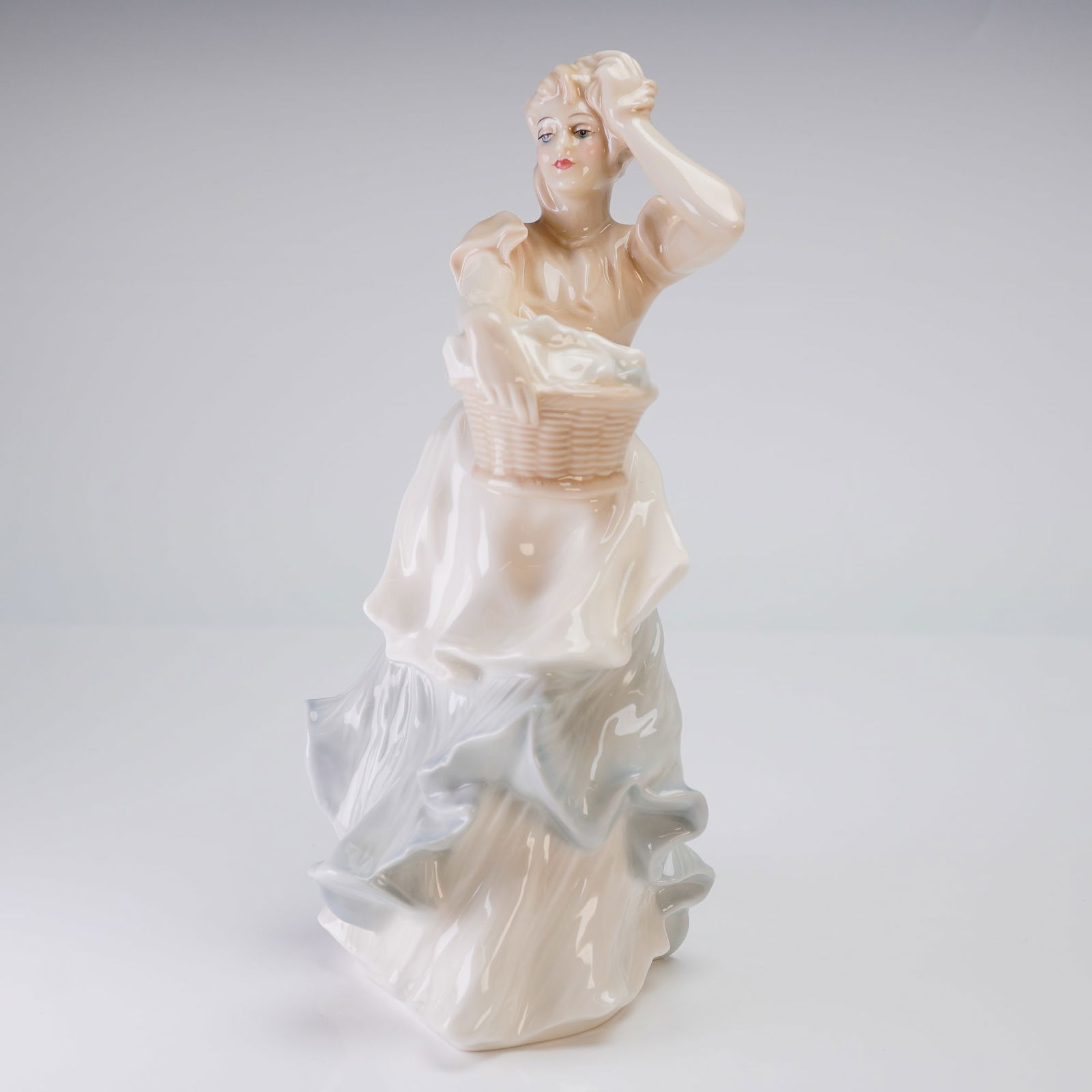 Royal Doulton Figurine, Reflections Breezy Day HN3162: The figure, modeled by Adrian Hughes, depicts a woman standing amidst wind-swept drapery, holding a basket in her left arm while her right hand is raised near her head, suggesting movement. The
