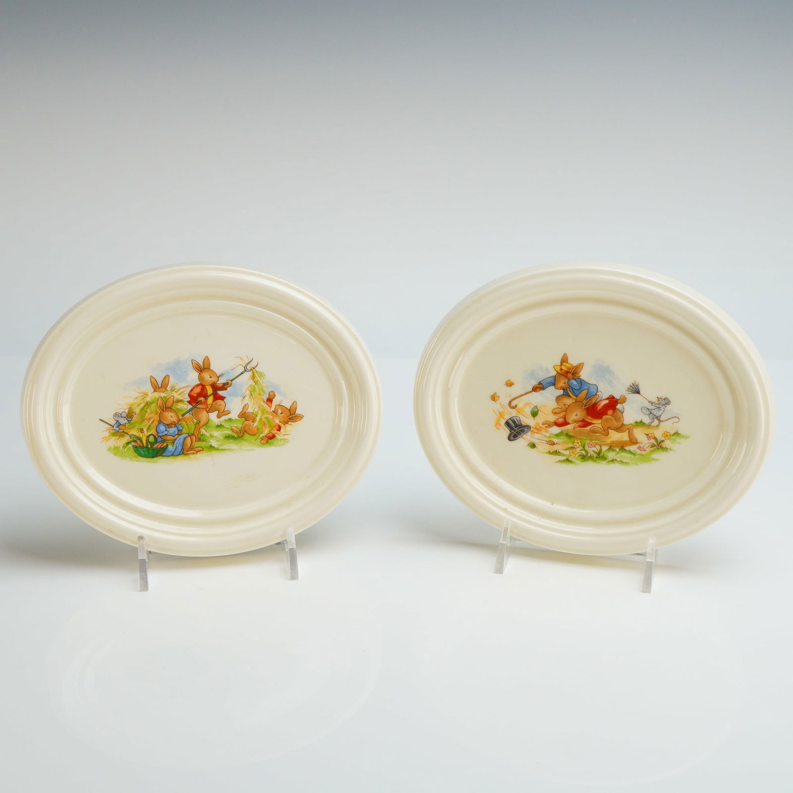 2pc Royal Doulton Bunnykins Wall Plaques: A pair of Royal Doulton Bunnykins wall plaques crafted from English fine bone china. These oval-shaped plaques feature transfer-printed illustrations of anthropomorphic rabbits. One plaque depicts a