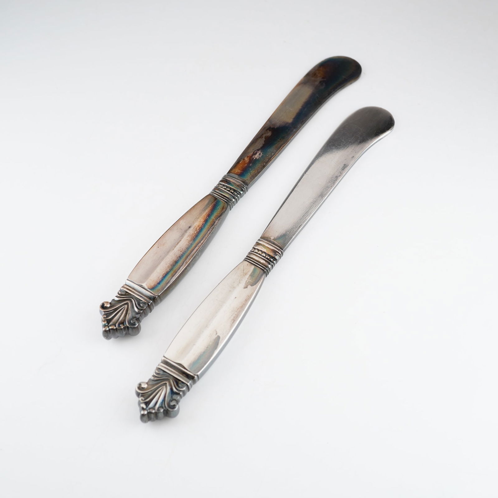2pc Georg Jensen Sterling Silver Butter Spreaders, Acanthus: Each piece features a handle with a decorative acorn-motif finial and a rounded, blunt-ended blade. The items are marked with the Georg Jensen dotted circle hallmark and stamped STERLING DENMARK on th
