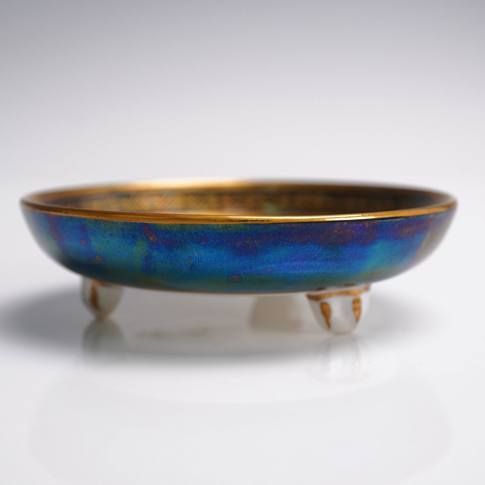 Wedgwood Fairyland Lustre Small Tri-Footed Bowl, Nizami Z5494: This small tri-footed bowl features a vibrant and iridescent moonlight blue ground under mother-of-pearl lustre. The interior is adorned with a finely detailed and multicolored scene inspired by the P
