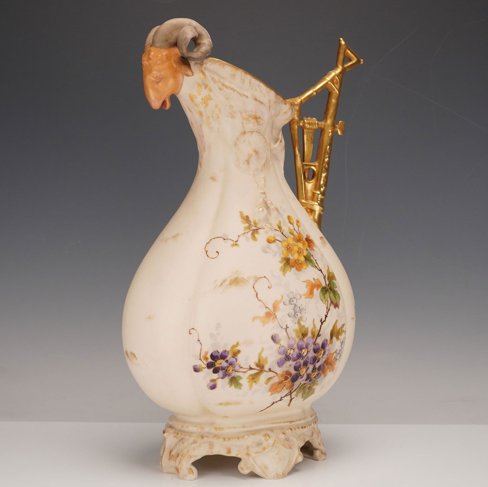 Alfred Stellmacher Turn-Teplitz Porcelain Floral and Ram Spout Ewer: A baluster-form, lobed body in an off-white glaze, decorated with hand-painted floral and vine motifs in purple, yellow, orange, and green, with gilt accents. The molded spout is formed as a ram's hea