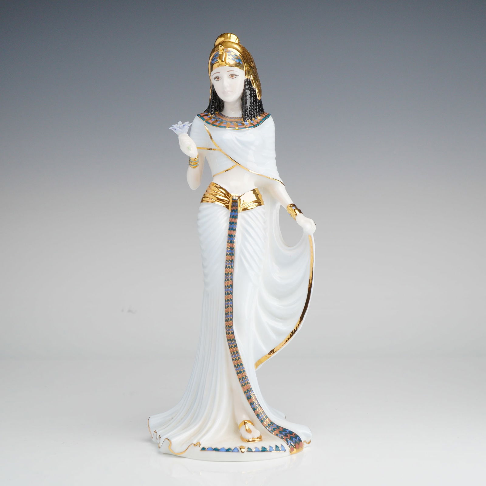Coalport Bone China Figurine, Cleopatra: This glazed bone china figure is modeled after Cleopatra, dressed in a flowing white top and skirt adorned with 22 carat gold detailing. She wears a gold headpiece and holds a blue flower. The undersi