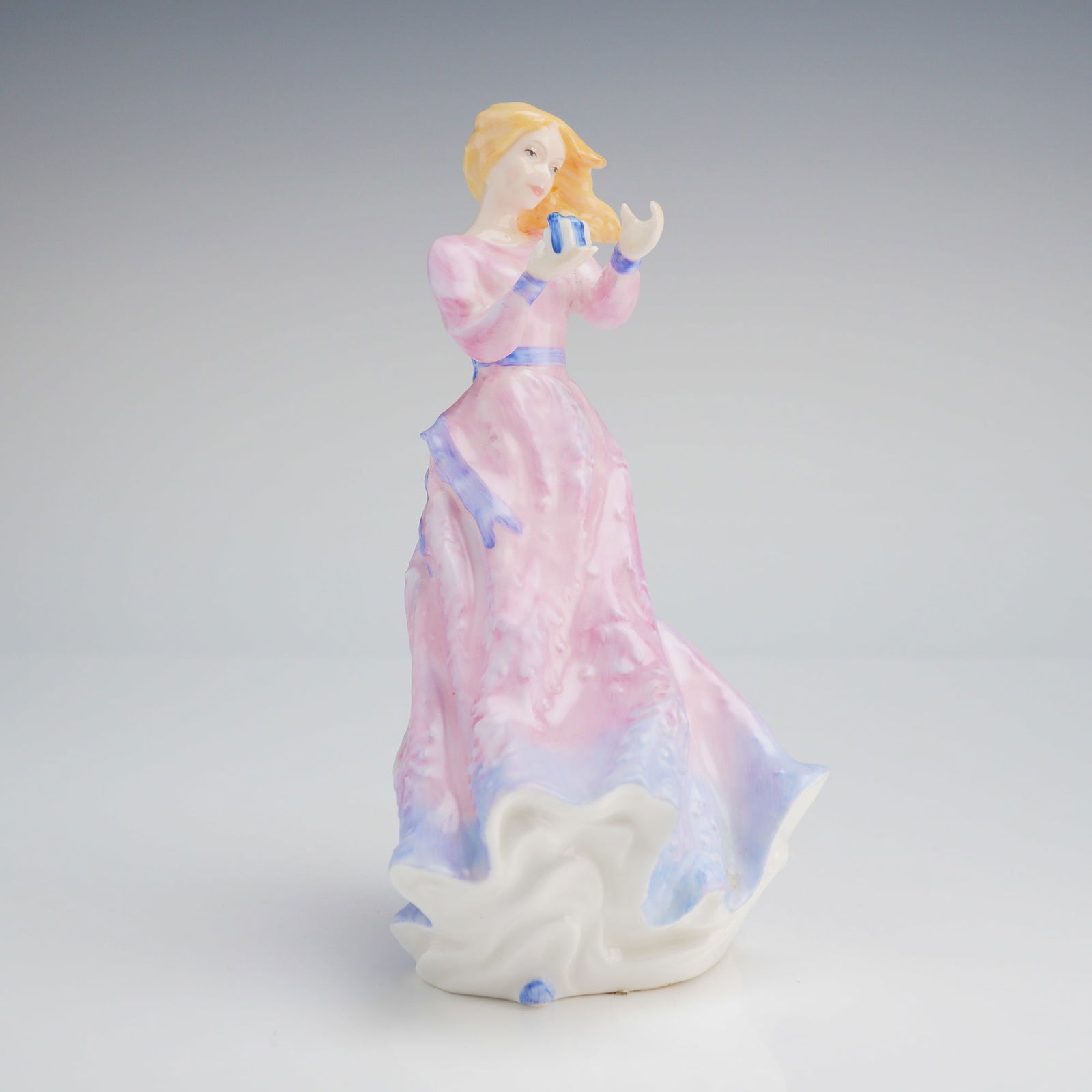 Royal Doulton Figurine, Liza Prototype: A Royal Doulton Liza-Prototype porcelain figurine featuring a woman in a flowing pink and blue dress holding a small blue object. Dimensions5.75"H Weight7.00oz