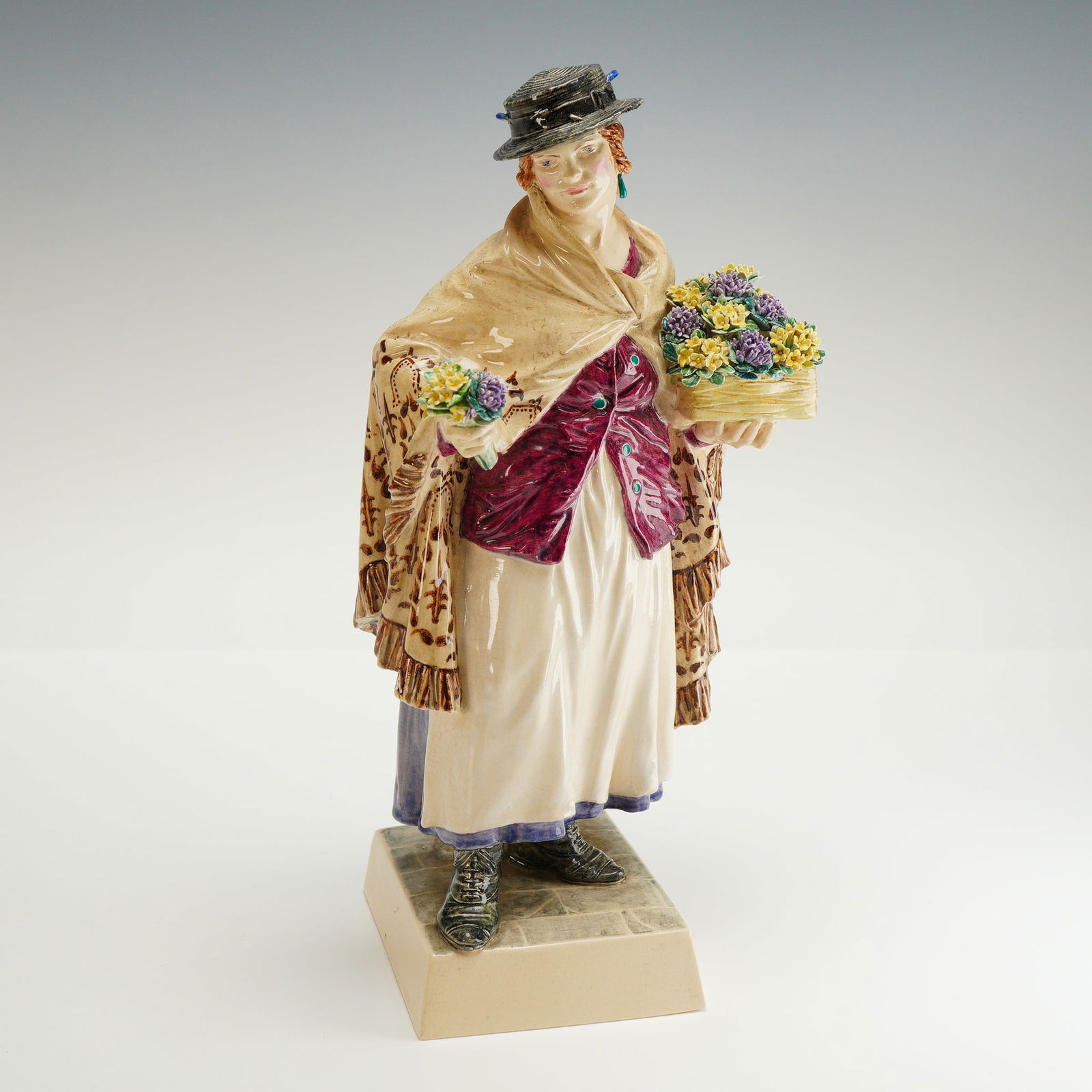 Charles Vyse Ceramic Figurine, The Shawl: A hand-painted ceramic figurine by Charles Vyse, depicting a flower selling woman holding a small bouquet of violet and yellow flowers in her right hand, and a flower box on her left arm. She is portr