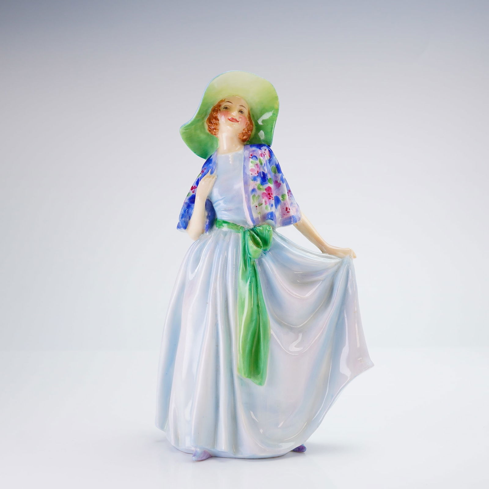 Royal Doulton Figurine Nadine HN1885: This Royal Doulton figurine is designed in an Art Deco style with a glossy glazed finish. The figure depicts a woman wearing a flowing light blue dress with a prominent green sash. Her attire includes