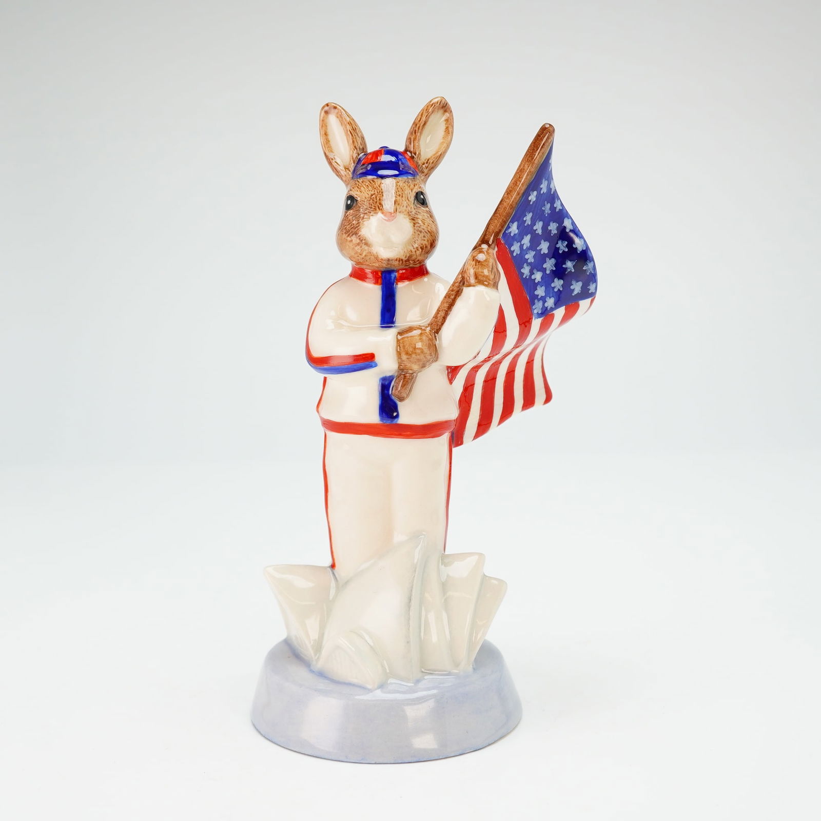 Royal Doulton Bunnykins Prototype Colorway Figurine, USA Athlete: A colorway exclusive variation of the Athlete Bunnykins Sydney 2000 (DB216), typically depicted representing England, but in this example featuring the flag of the United States. The figure is dressed