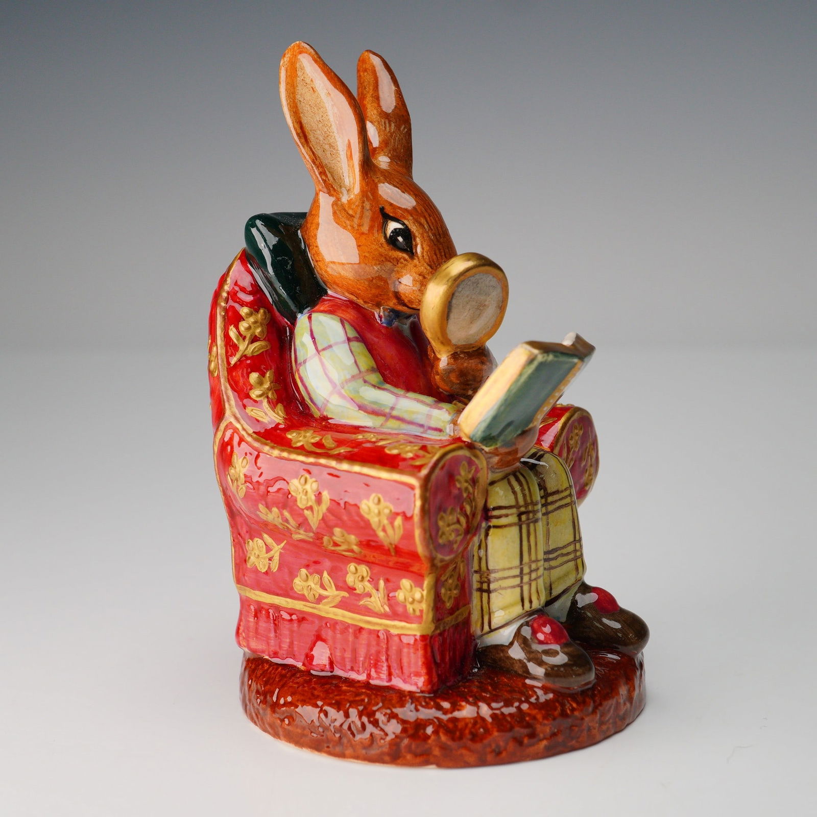 Royal Doulton Sample Prototype Bunnykins, The Collector: A colorway of The Collector DB54. Modeled with a rose color vest and plaid shirt and pants. Bunnykins sits in a red and gold floral armchair. Sample backstamp. Royal Doulton backstamp. Dimensions2.75"