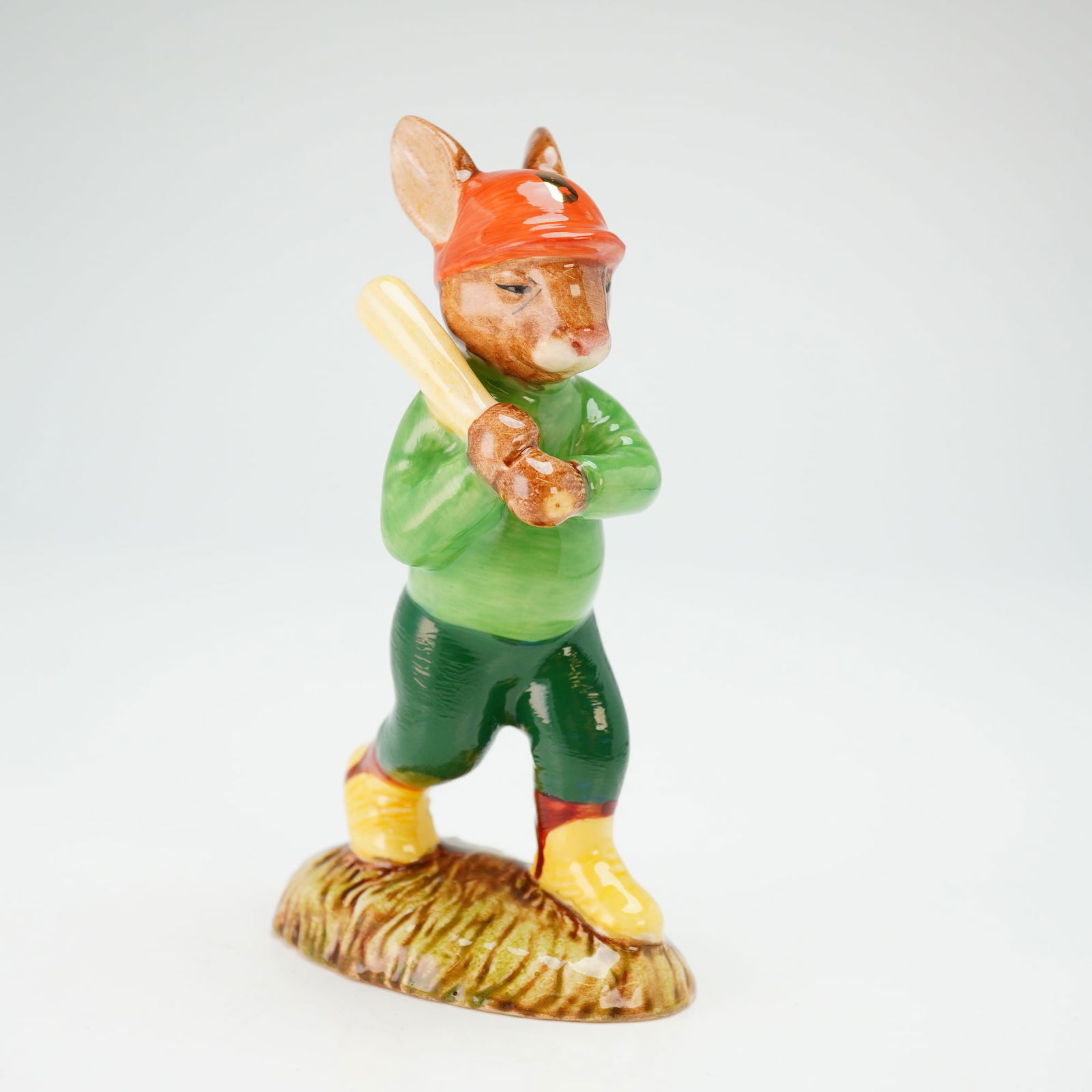Royal Doulton Prototype Artist Copy Bunnykins Figurine, Home Run DB43: A porcelain figure depicting a bunny holding a baseball bat in a swinging pose, dressed in a light green sweater, dark green pants, yellow shoes, and an orange hat marked with a gold letter "B." The u