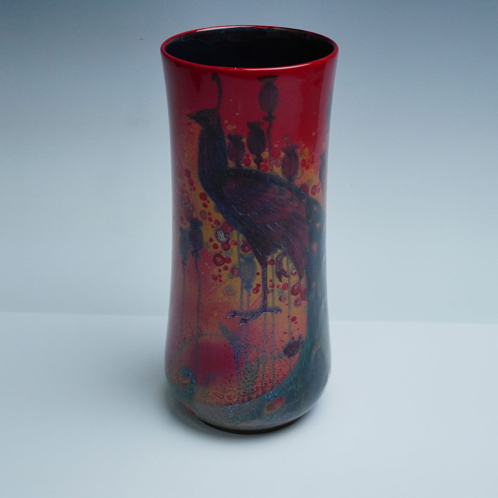 Royal Doulton Flambe Sung Peacock Vase: Royal Doulton flambe vase from the Sung series featuring a stylized depiction of a peacock among tall seed pods. The cylindrical vase displays a vibrant red flambe glaze with transitions to orange and