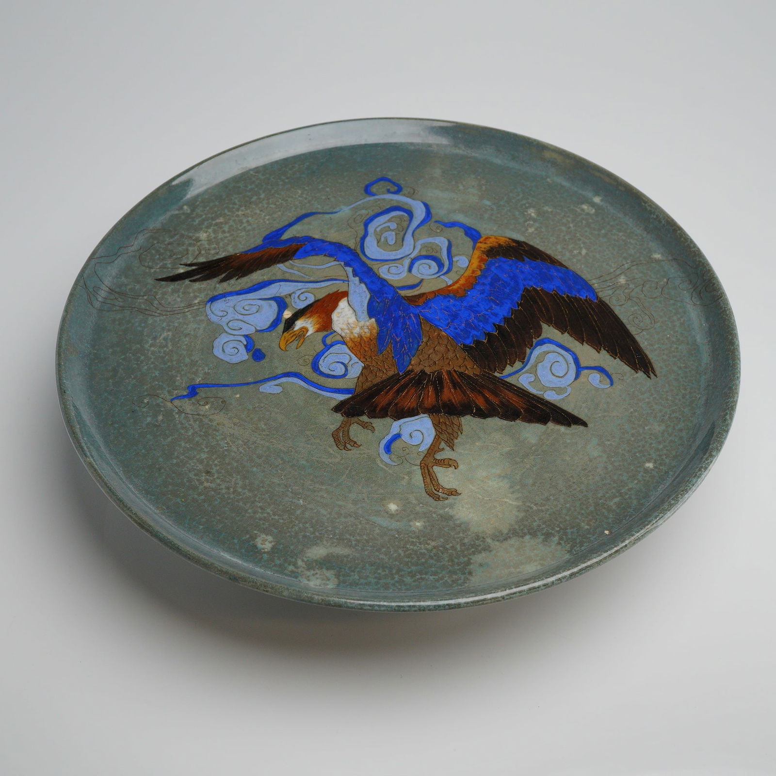 Royal Doulton Titanian Eagle Footed Plate: A round plate with a footed base, finished in a mottled green gray glaze. The center features a hand painted eagle in mid flight, surrounded by swirling clouds, executed in shades of light blue, royal