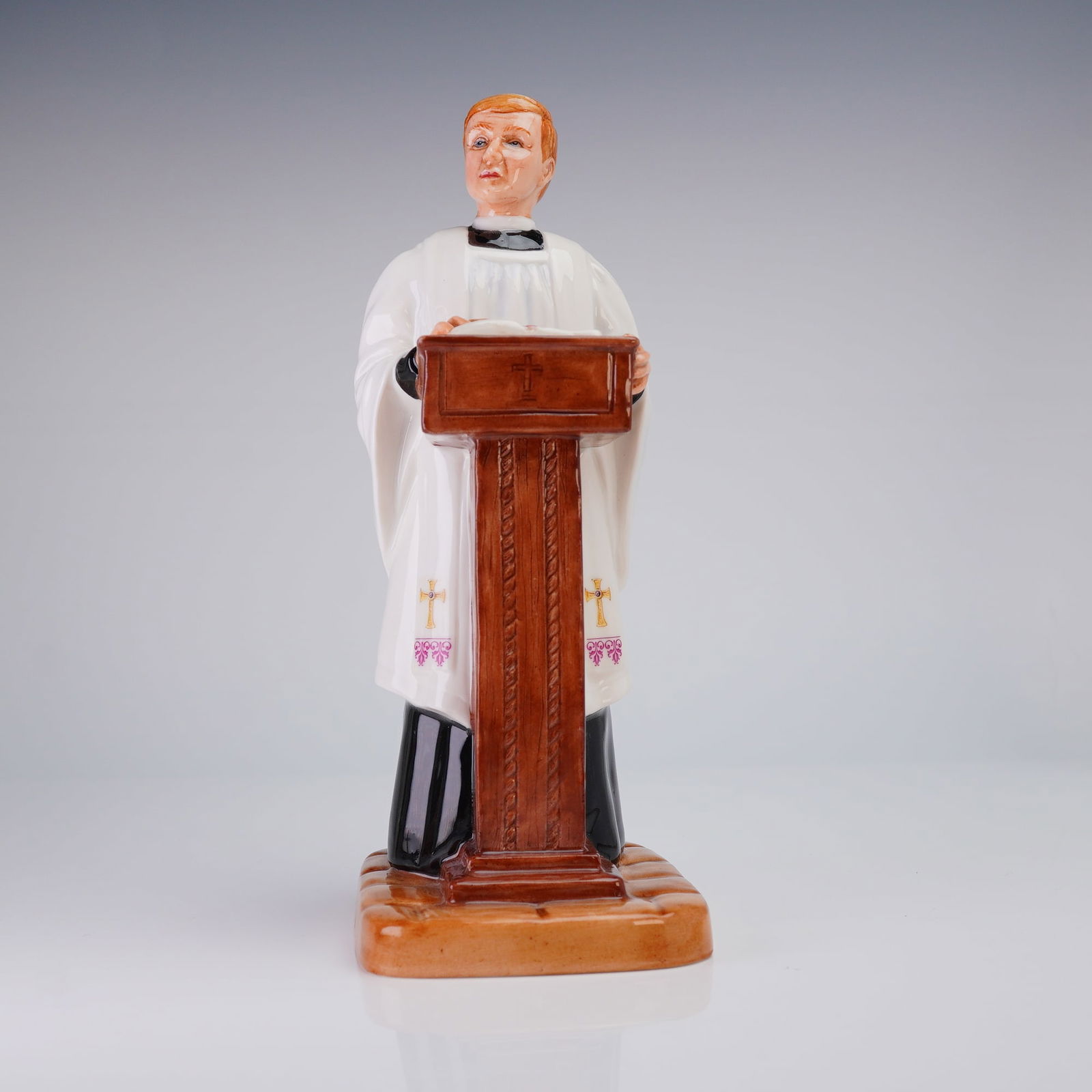 Royal Doulton Prototype Figurine, Vicar: English porcelain figurine of a vicar at a pulpit with an open book, finished in glaze and wearing a black cassock, white surplice, and white stole with gilt crosses and pink floral accents. Underside