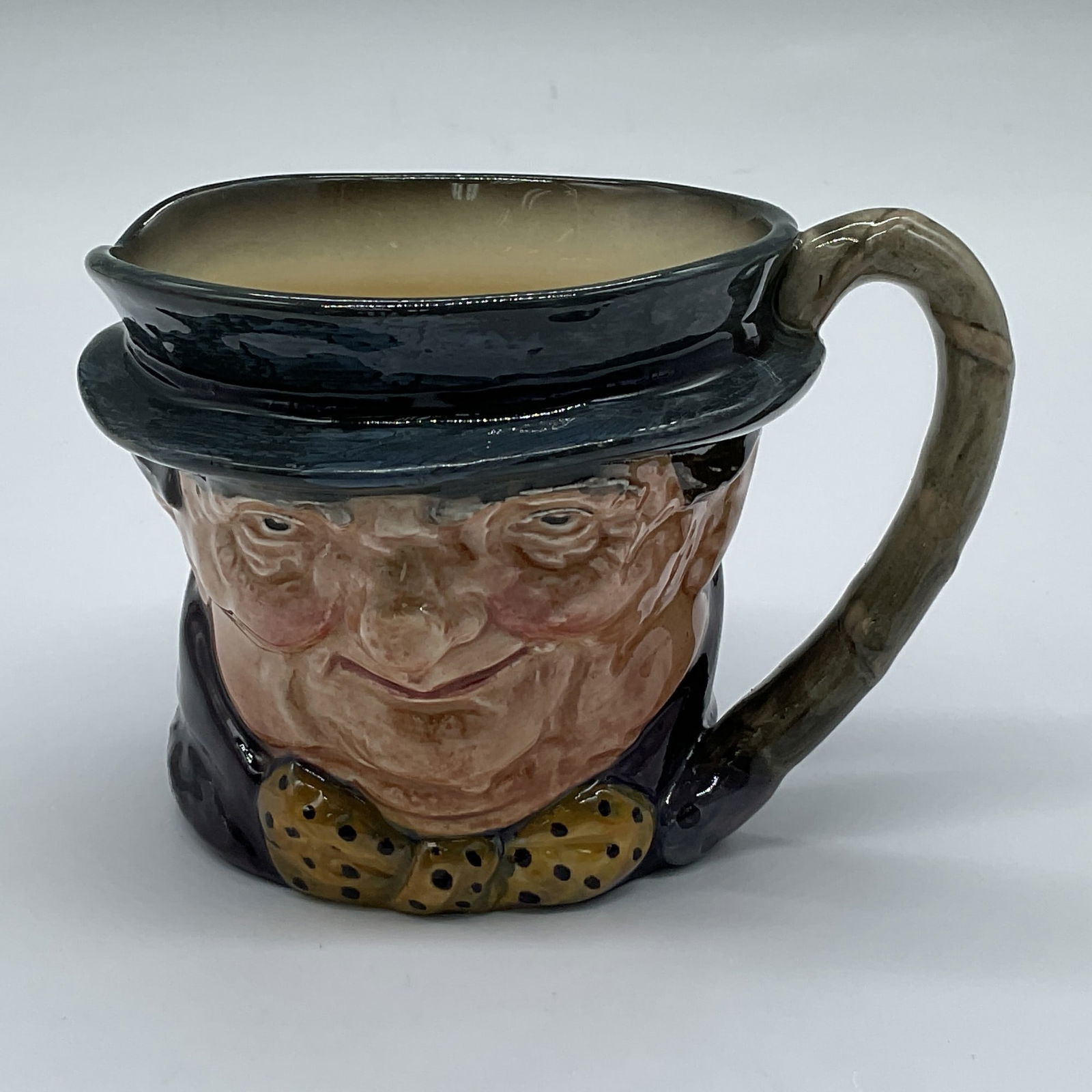 Royal Doulton Colorway Small Character Jug, Tony Weller: D5530. Hand painted Charles Dickens character figural jug, depicted in a gray-blue hat with a purple band, and a dark purple jacket with a mustard-yellow bowtie with black spots. Gray handle. Royal Do