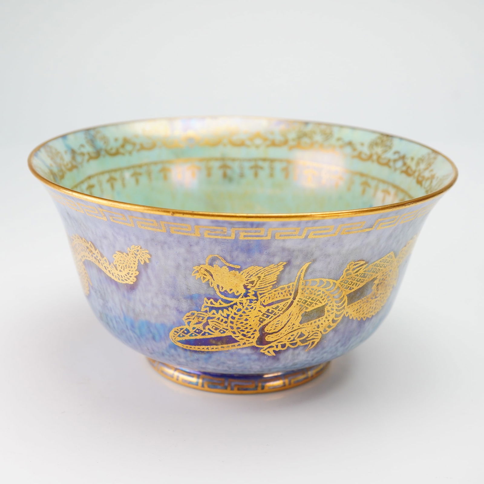 Wedgwood Fairyland Lustre Dragon Bowl: This footed ceramic vessel features an iridescent mottled blue and purple glazed exterior decorated with gilded dragons and a Greek key border at the rim. The interior is finished in a pale green and