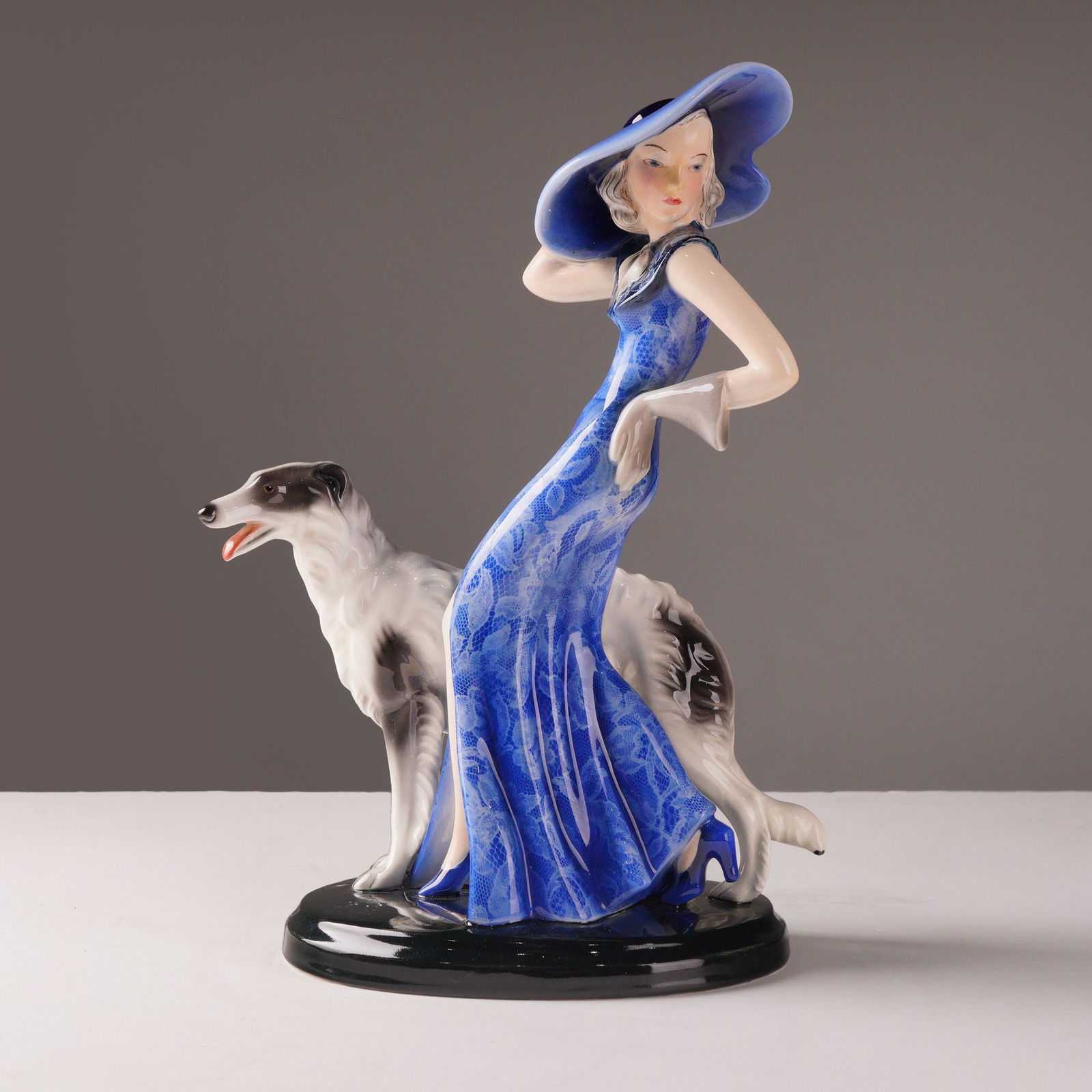 Goldscheider by Claire Herczeg-Weiss Sculpture, Lady with Borzoi: A high-gloss ceramic Art Deco figure designed by Claire Herczeg-Weiss ("Claire Herczeg" on the underside). The piece portrays an elegant, woman in a sweeping blue gown with a floral lace overlay, comp
