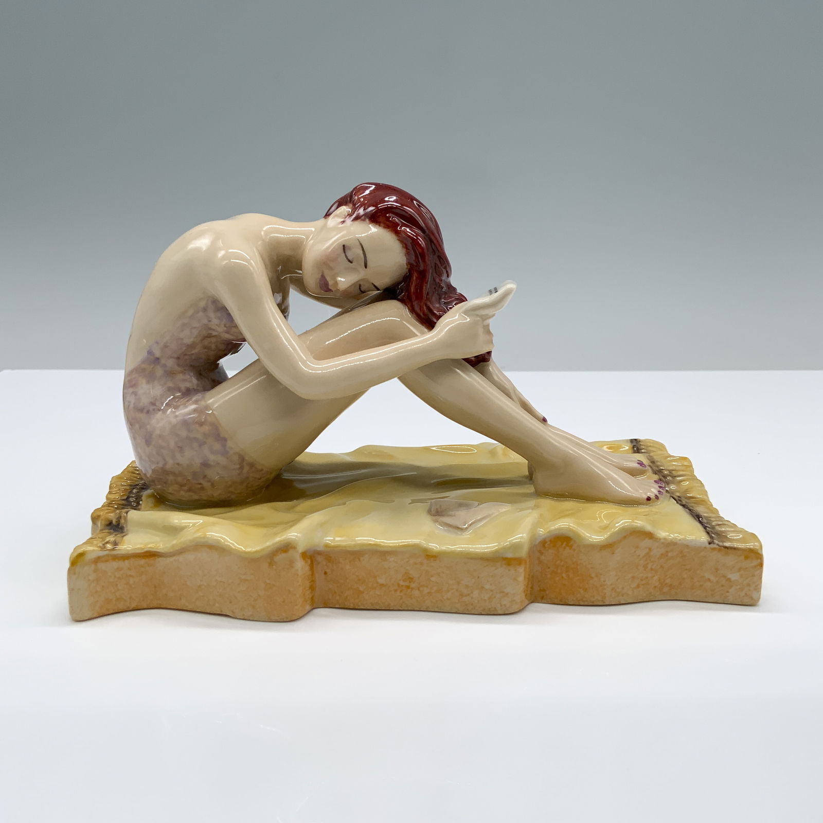 Peggy Davies Ceramics Figurine, Valentine: Hand painted porcelain of a seated woman with red hair, delicately combing her hair while resting on a yellow blanket. Peggy Davies Ceramic backstamp. Pin-Up Parade backstamp. Dimensions6.75"L x 3.75"