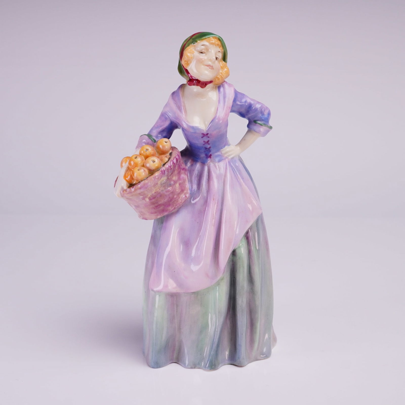 Royal Doulton Figurine Orange Seller HN1325: The single female figure is rendered in polychrome enamels, characteristic of mid-20th century English pottery figures. She wears a long, layered skirt in blended tones of green and lilac/purple, a pu
