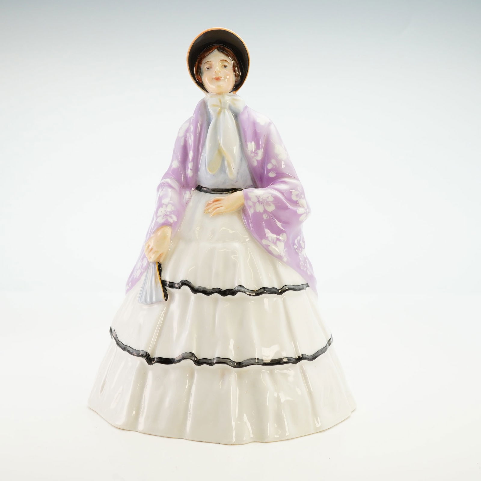 Royal Doulton Colorway Figurine, Lilac Shawl HN44: A hand-painted figure of a woman wearing a white layered skirt with black outlining, a light blue top, and a peach and black bonnet. A lilac shawl decorated with white flowers is draped over her shoul