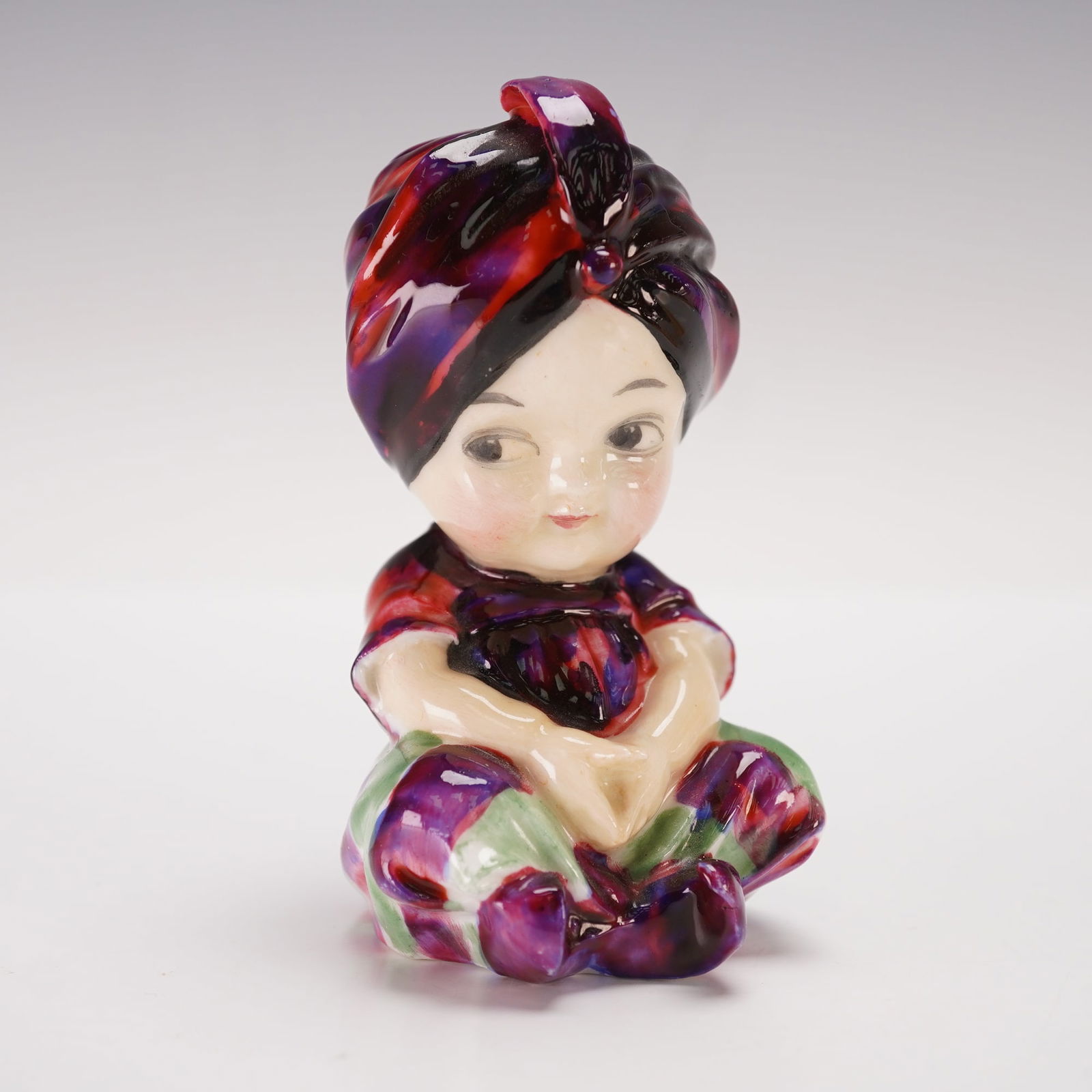 Rare Royal Doulton Figurine, Boy with Turban HN1212: This rare figurine depicts a young boy seated with his legs crossed, wearing a dark turban in shades of red and purple. His shirt is a deep, rich color, complemented by striped pants in green and redd