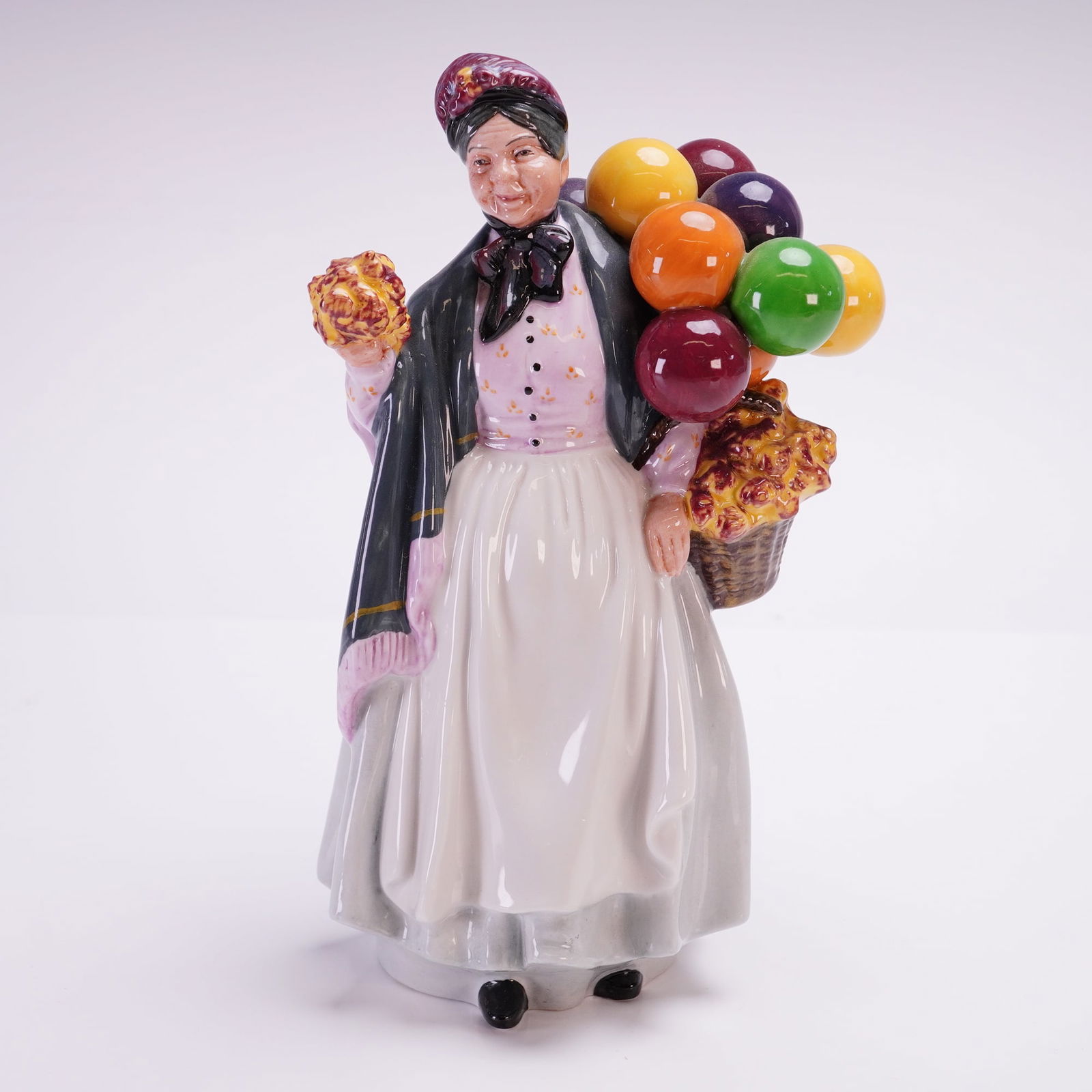 Royal Doulton Figurine Biddy Penny Farthing HN1843: This hand-painted and hand-decorated piece depicts an elderly woman in traditional Victorian-style attire, including a dark shawl with pink fringed edging, a white apron over a gray skirt, and a patte