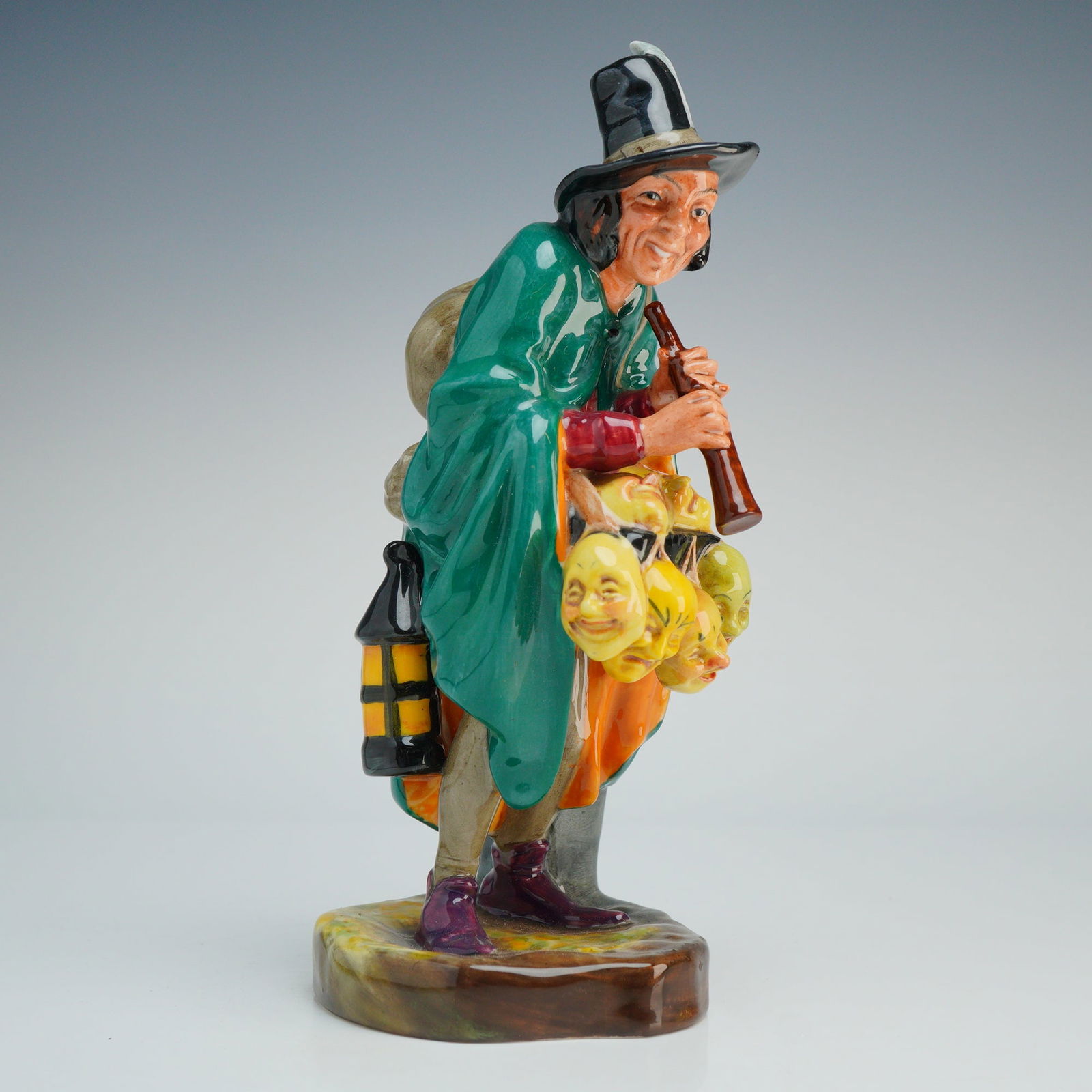 Royal Doulton Figurine, Mask Seller HN2103: This Royal Doulton glazed figurine is a hand-painted porcelain piece. The figure depicts a male character wearing a green cloak and a tall black hat featuring a white feather. He is shown playing a br