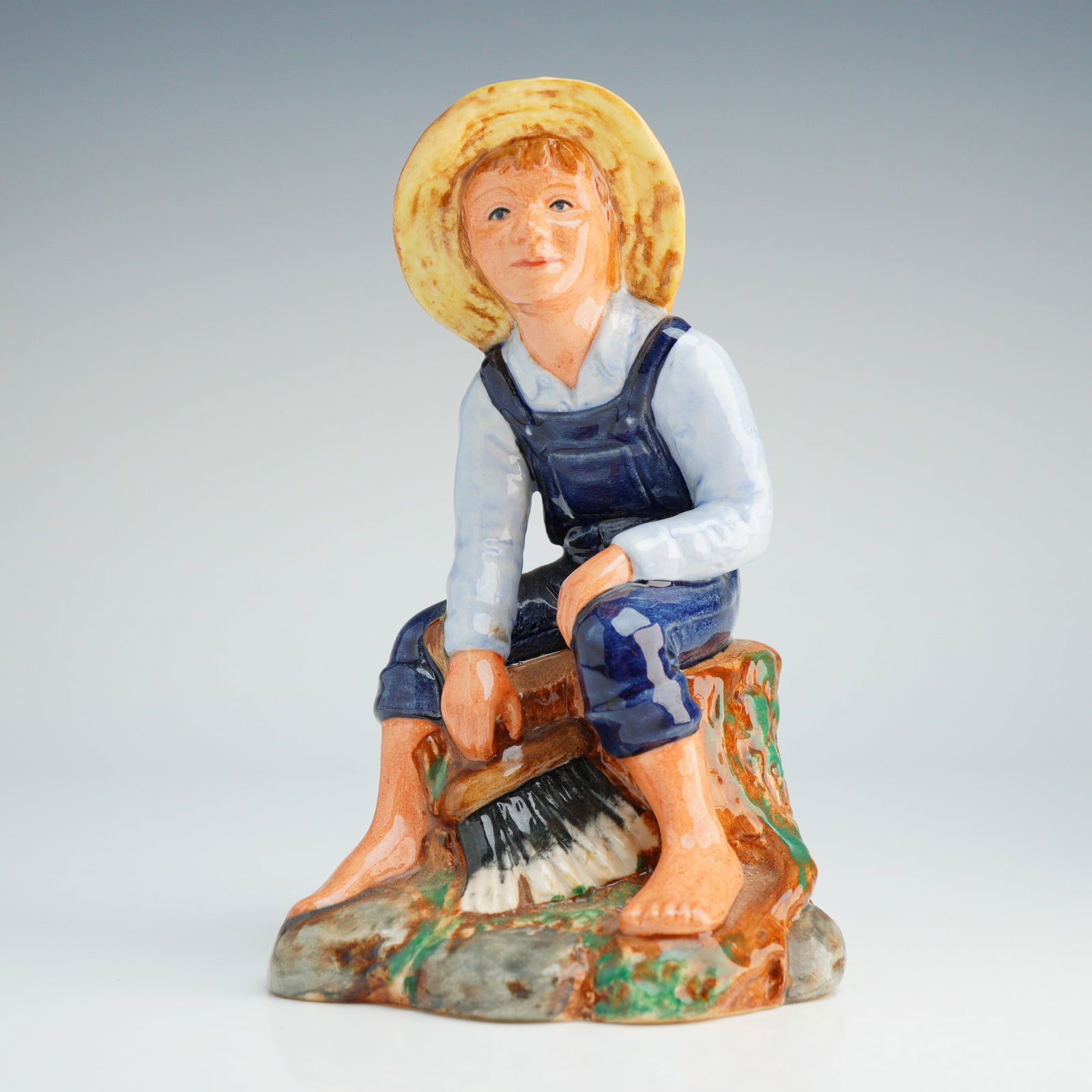 Royal Doulton Figurine, Tom Sawyer HN2926: This polychrome glazed ceramic figurine, produced by Royal Doulton, depicts the literary character Tom Sawyer. The barefoot boy is shown sitting on a tree stump base, wearing a straw hat, a light-colo