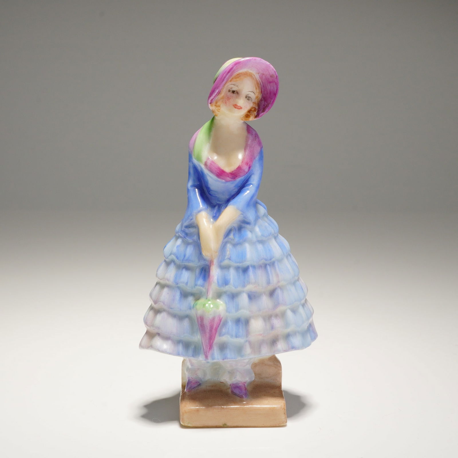 Royal Doulton Miniature Figurine Priscilla M14: A single Priscilla M14 Royal Doulton Figurine made of bone china depicts a woman in a ruffled blue and pink dress holding an umbrella, marked on the base with the Royal Doulton logo and model number.