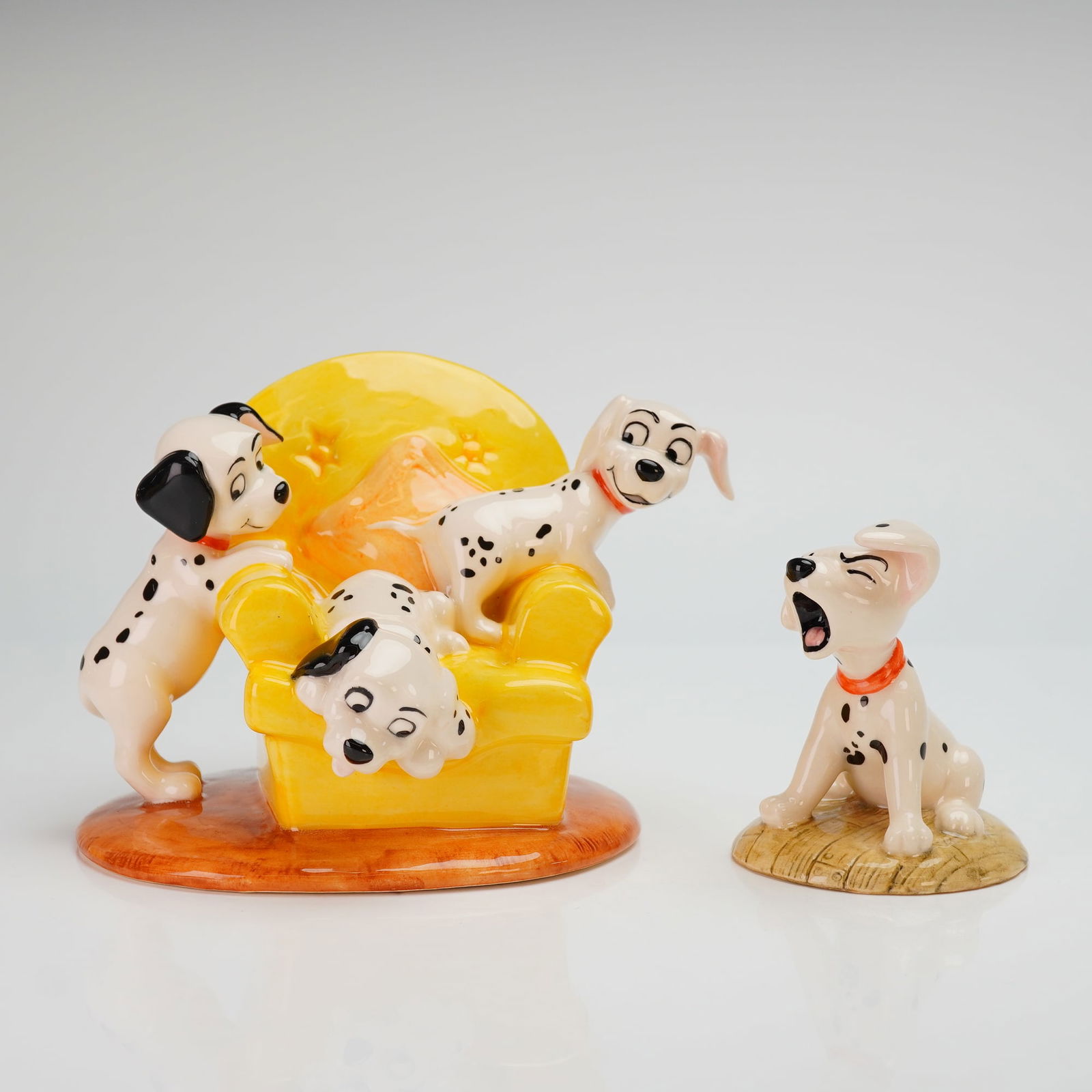 Two Royal Doulton Disney 101 Dalmatians Figurines, DM11 and DM02: The first piece, titled Pups in the Armchair (model DM 11), depicts a group of puppies playfully interacting with a bright yellow upholstered armchair on a circular base. The second figurine, Penny (m