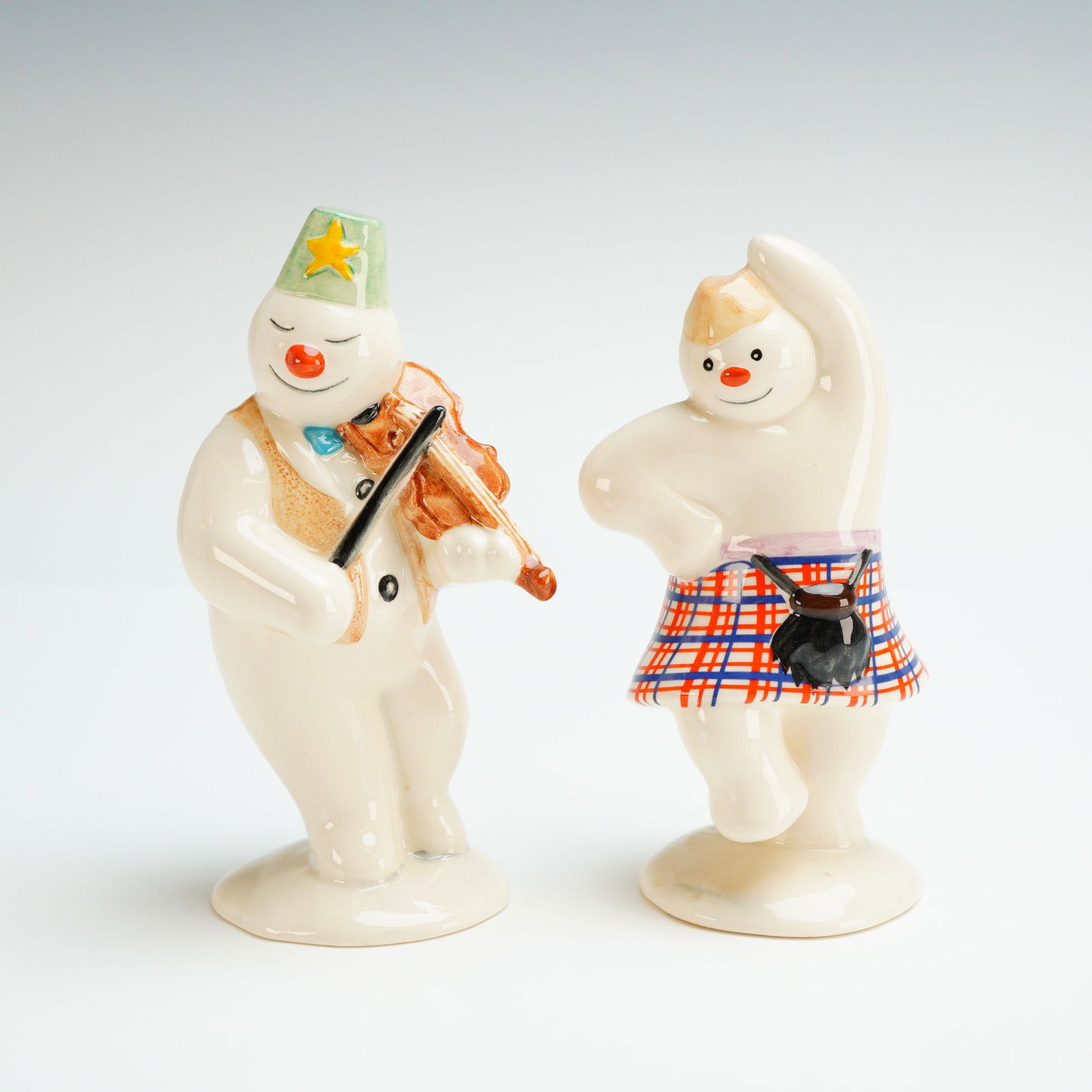 Two Royal Doulton Snowman Figurines, DS7 and DS11: A pair of Royal Doulton featuring the Highland Snowman (DS 7) and the Violinist Snowman (DS 11). These glazed ceramic figures are based on the character created by Raymond Briggs and were produced in