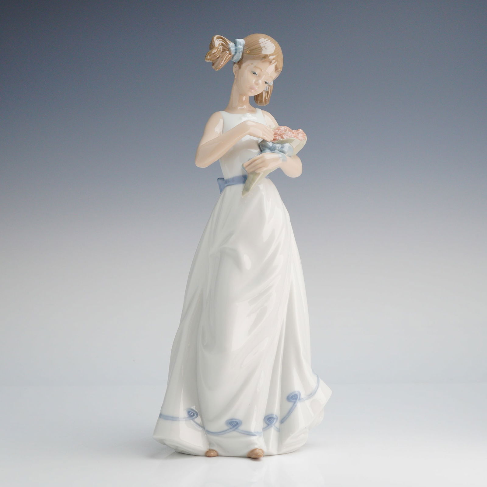 NAO by Lladro Porcelain Figurine, Girl with Holding a Bouquet: A NAO by Lladro glazed porcelain figurine of a girl with pigtails holding a bouquet. The figure is depicted in a standing pose, wearing a sleeveless white gown with a blue sash and a scrolled blue pat
