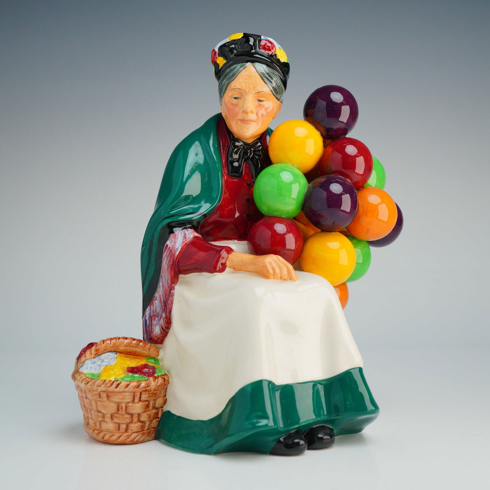 Royal Doulton Figurine, Old Balloon Seller HN1315: The piece depicts a seated elderly woman wearing a green shawl with a red and white patterned fringe, a red blouse, and a white apron. She is shown holding a large bunch of multi-colored balloons in v